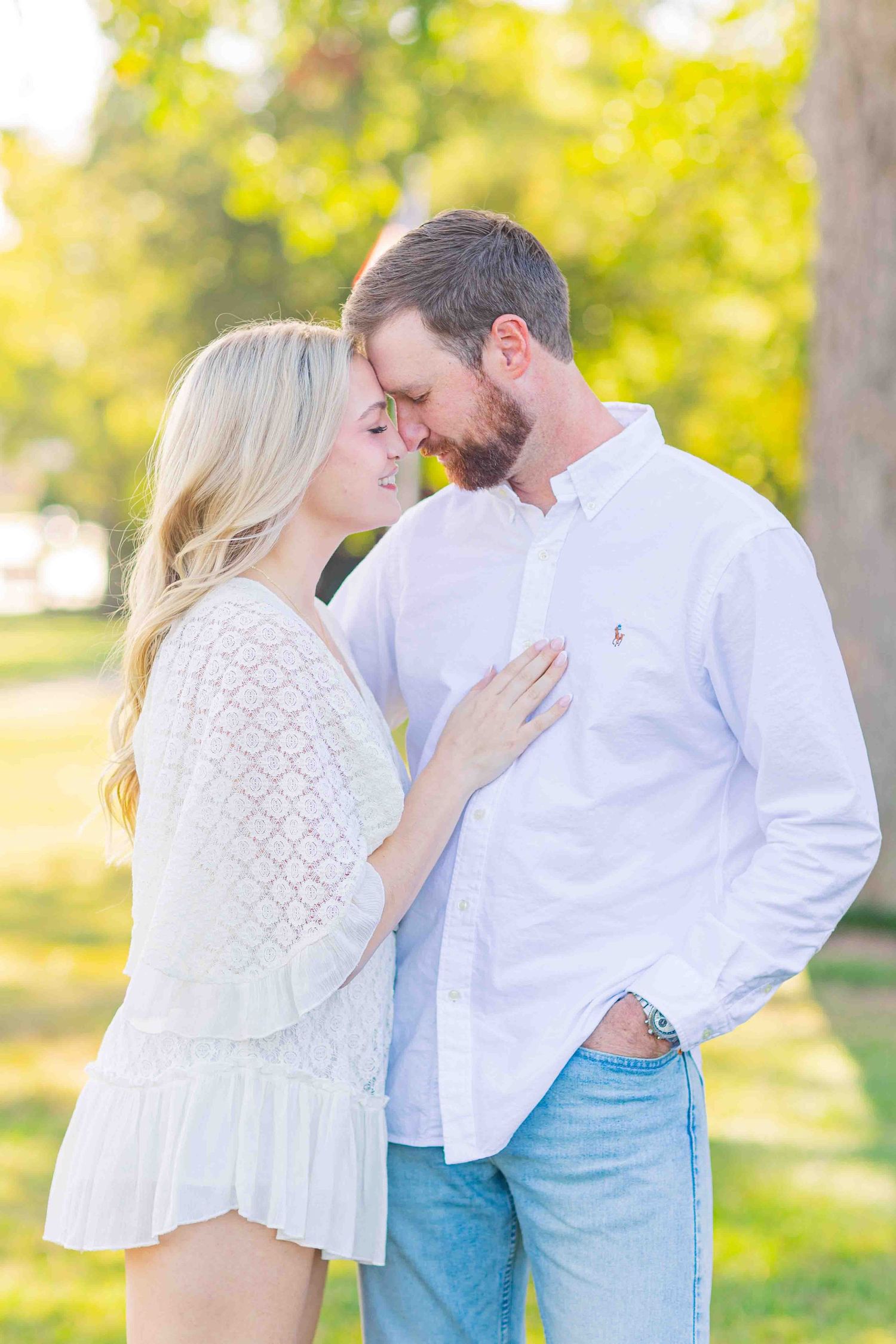 Couple sharing a tender moment in a sunlit park, woman in white dress touching mans chest.