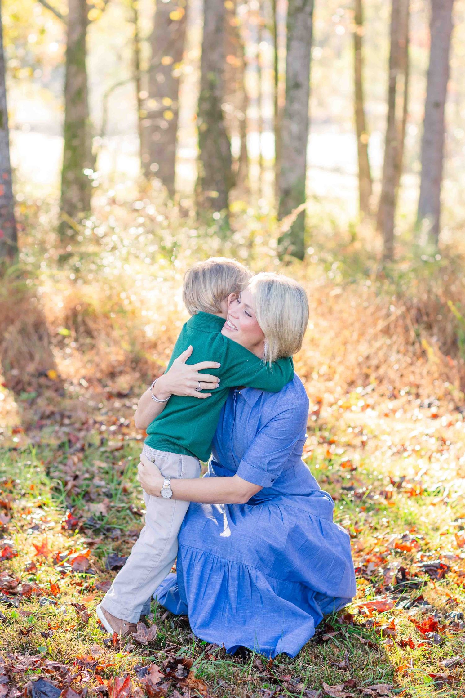 Blonde mother hugging and kissing her young son in a sunlit autumn woodland.