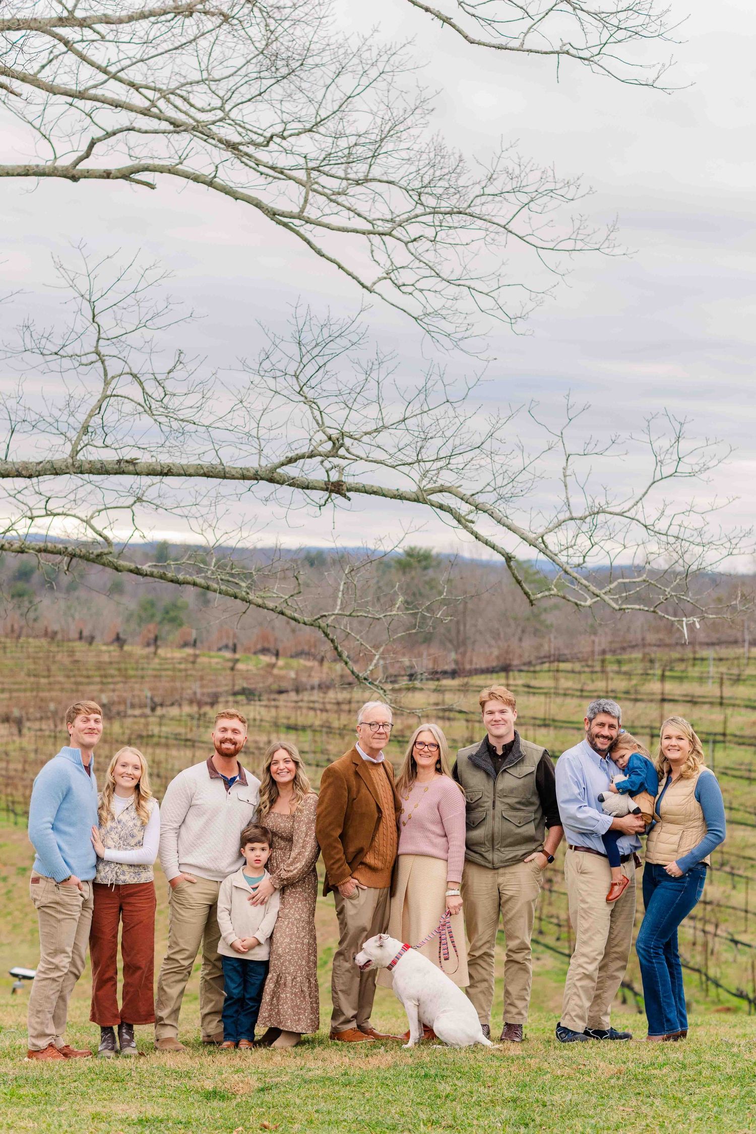Large extended family of ten adults and children posing outdoors at a winter vineyard with a dog.