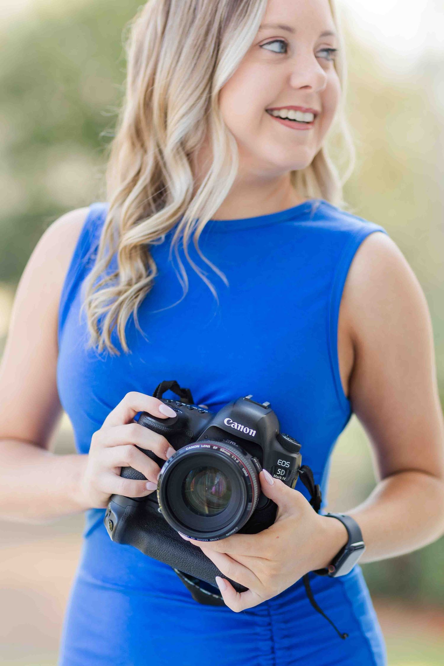 Smiling blonde female photographer in blue sleeveless dress holding a Canon EOS DSLR camera outdoors.