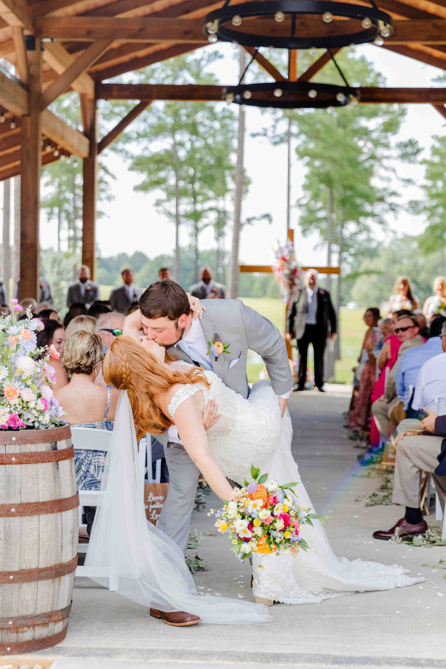 Bride and groom share a romantic dip kiss at the altar during an outdoor rustic barn wedding ceremony with colorful flowers.