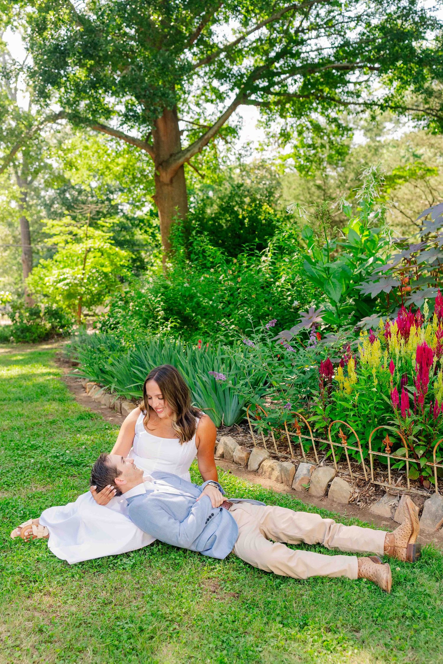 Couple relaxing on lush green grass in garden, woman in white dress sitting while man lies with head in her lap.