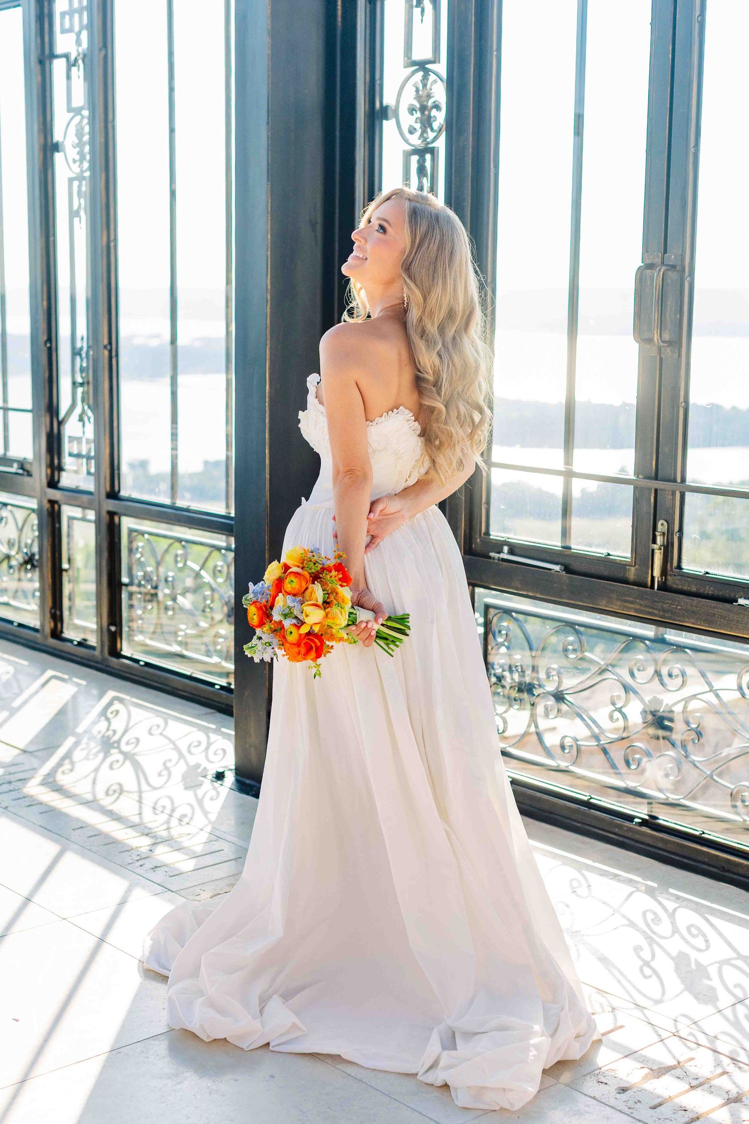 Blonde bride in strapless white gown holds colorful bouquet by ornate balcony windows with scenic view.