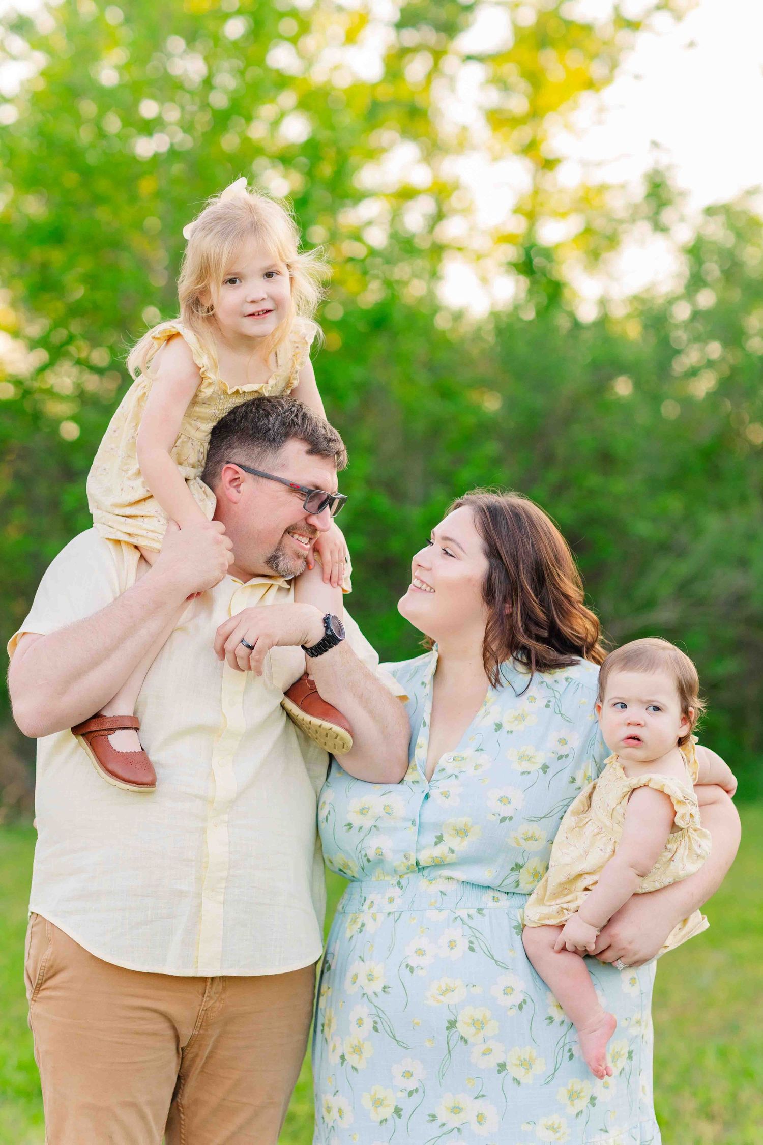 Happy family with dad carrying toddler on shoulders and mom holding baby in sunny green field.