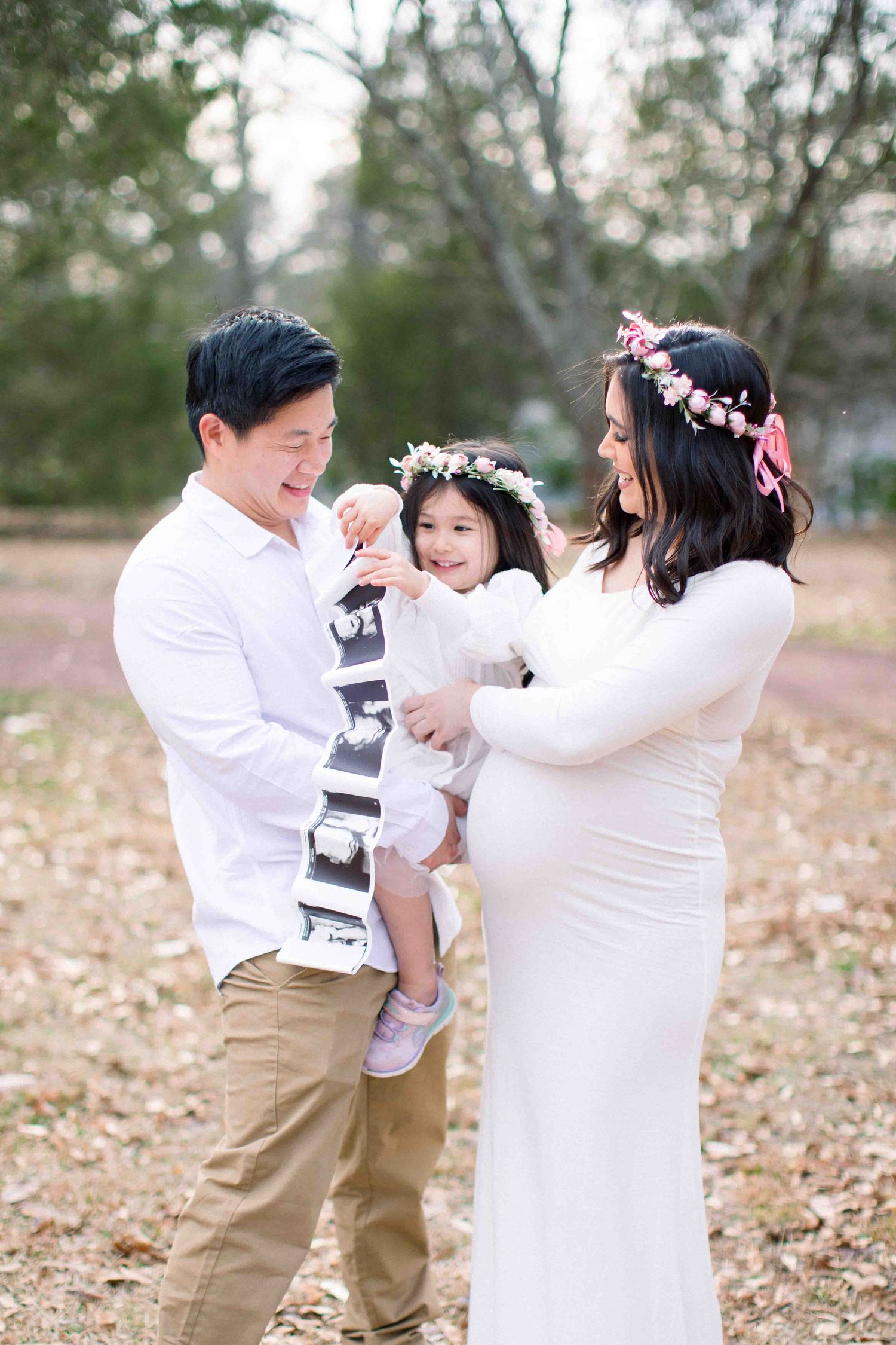 Expecting parents with toddler daughter holding ultrasound photos outdoors in white outfits.