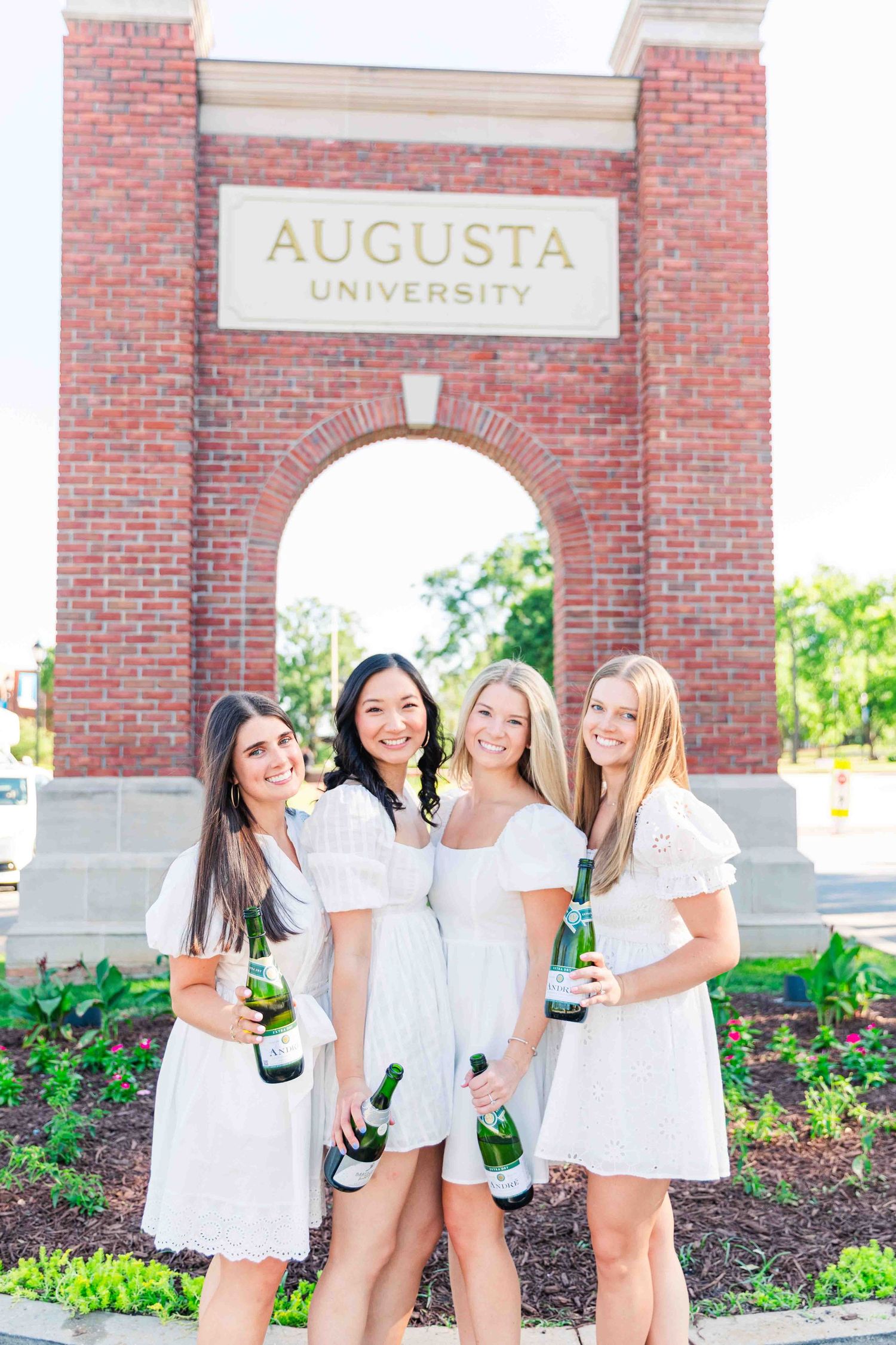 Four women in white dresses celebrate with champagne bottles at Augusta University arch.