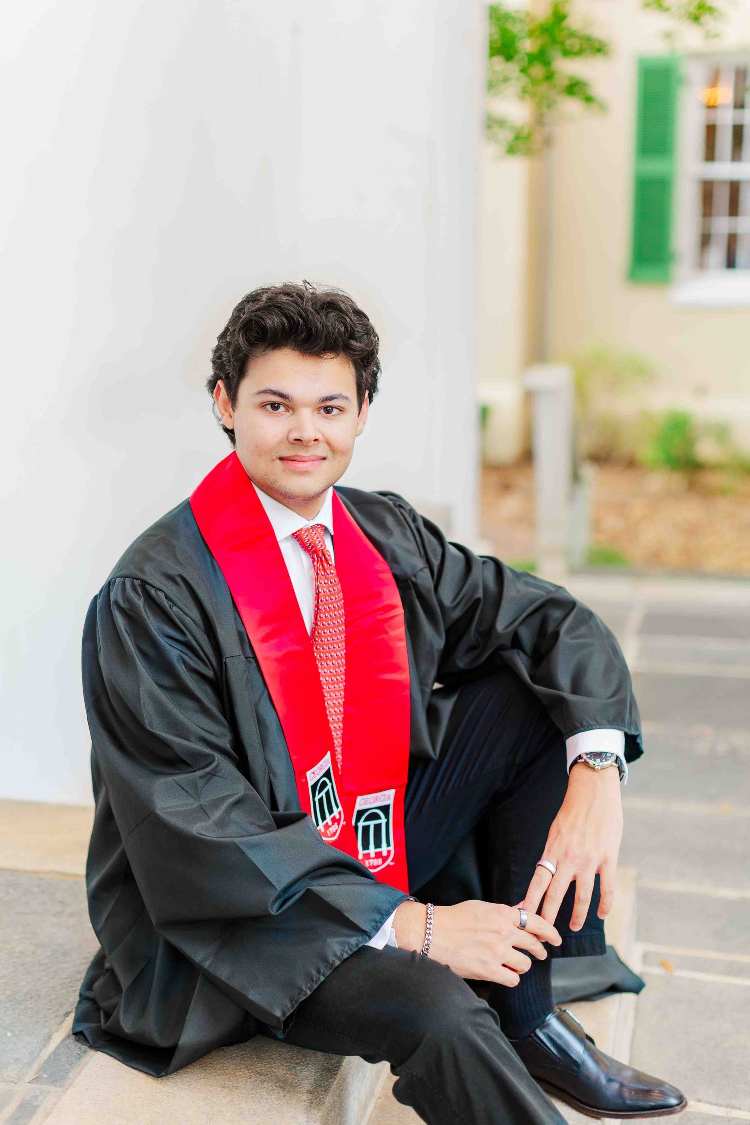 Smiling graduate in black gown and red stole sitting on steps outdoors for senior graduation photos.
