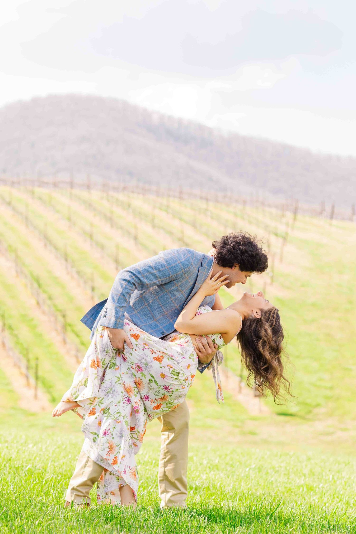 Man dipping woman for a romantic kiss on green grass with vineyard rows and mountain in background.
