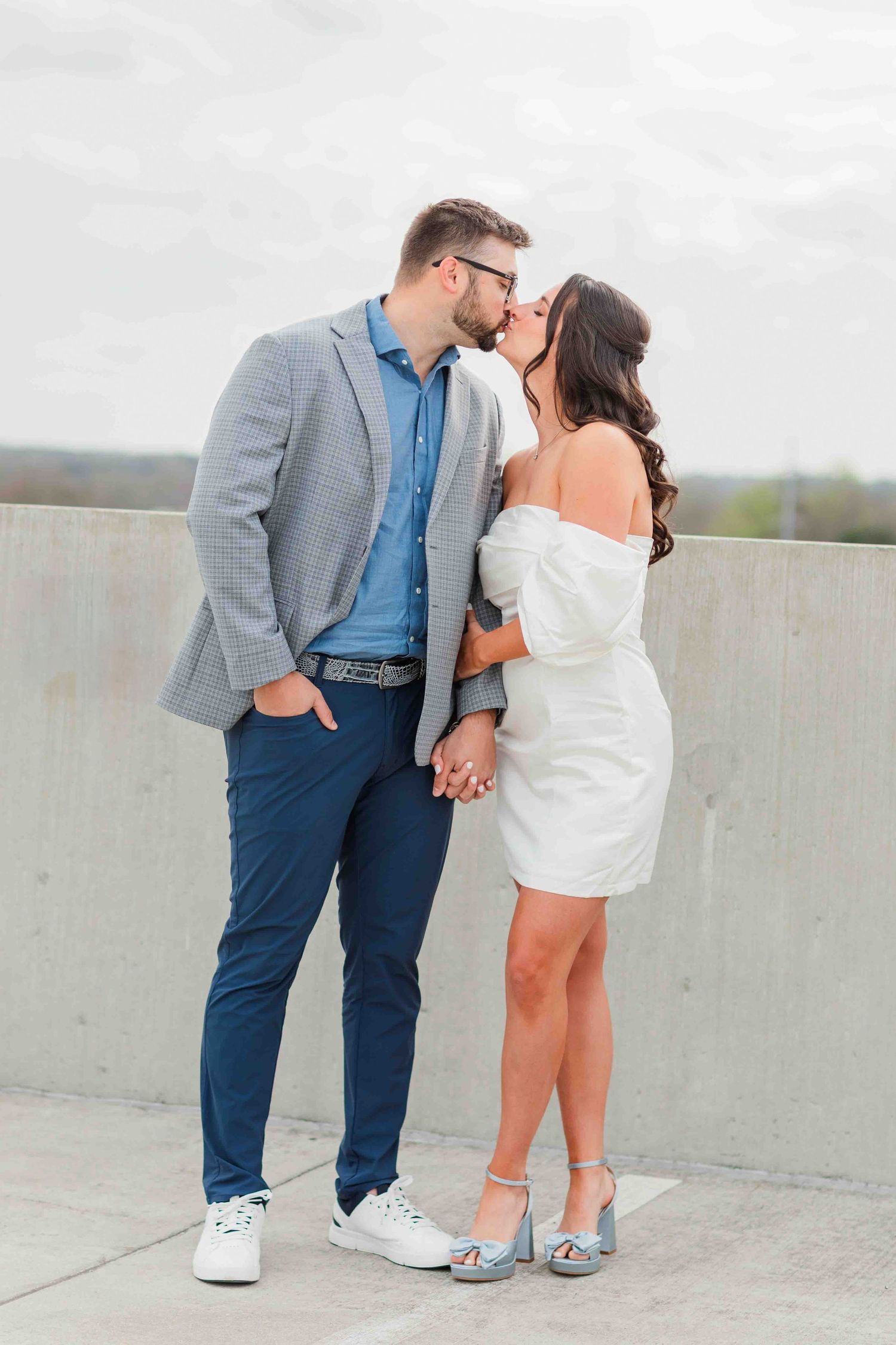 Couple kissing on rooftop, man in gray blazer and blue pants, woman in white mini dress and heels.