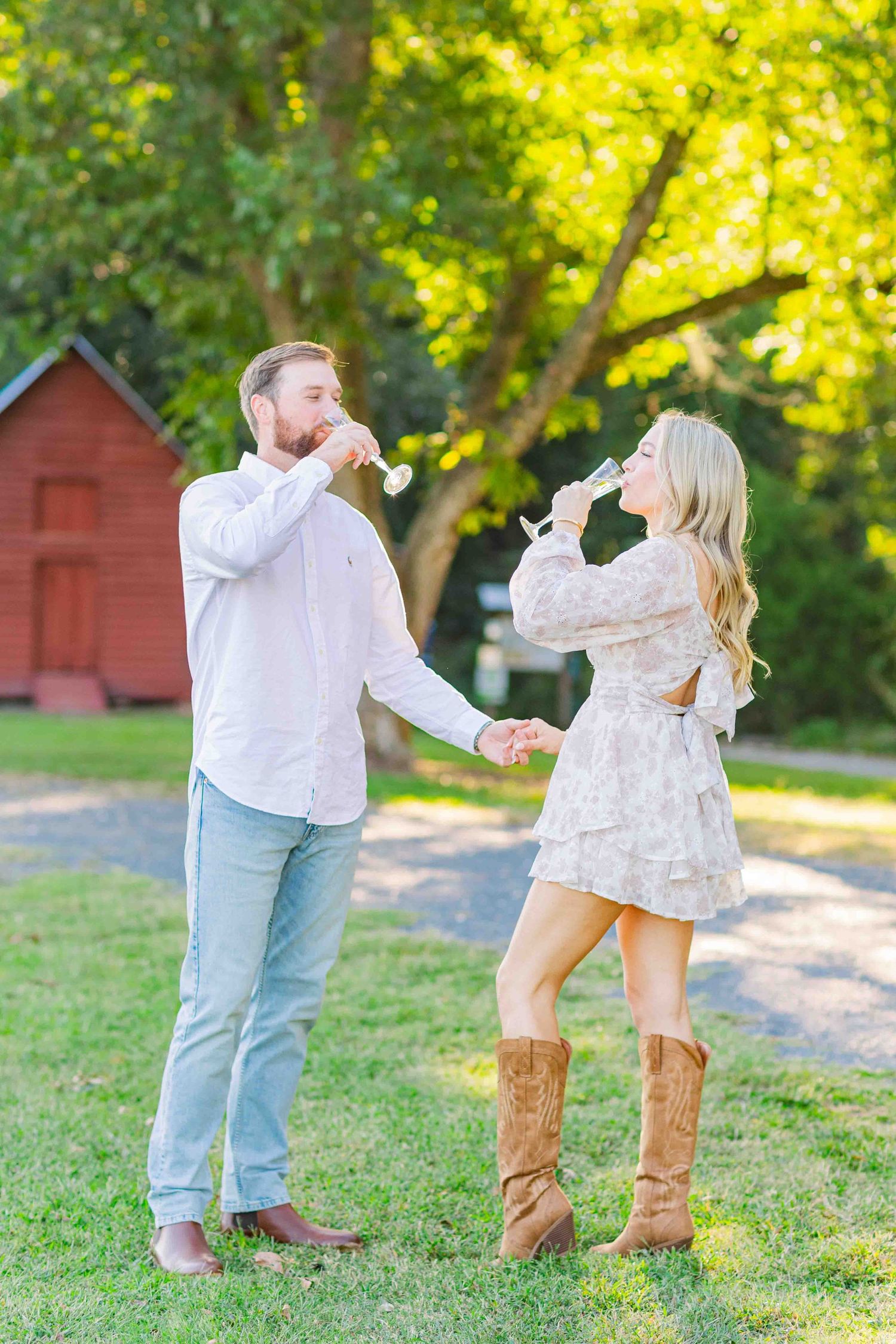 Couple celebrating engagement by drinking champagne while holding hands on a sunny green lawn.