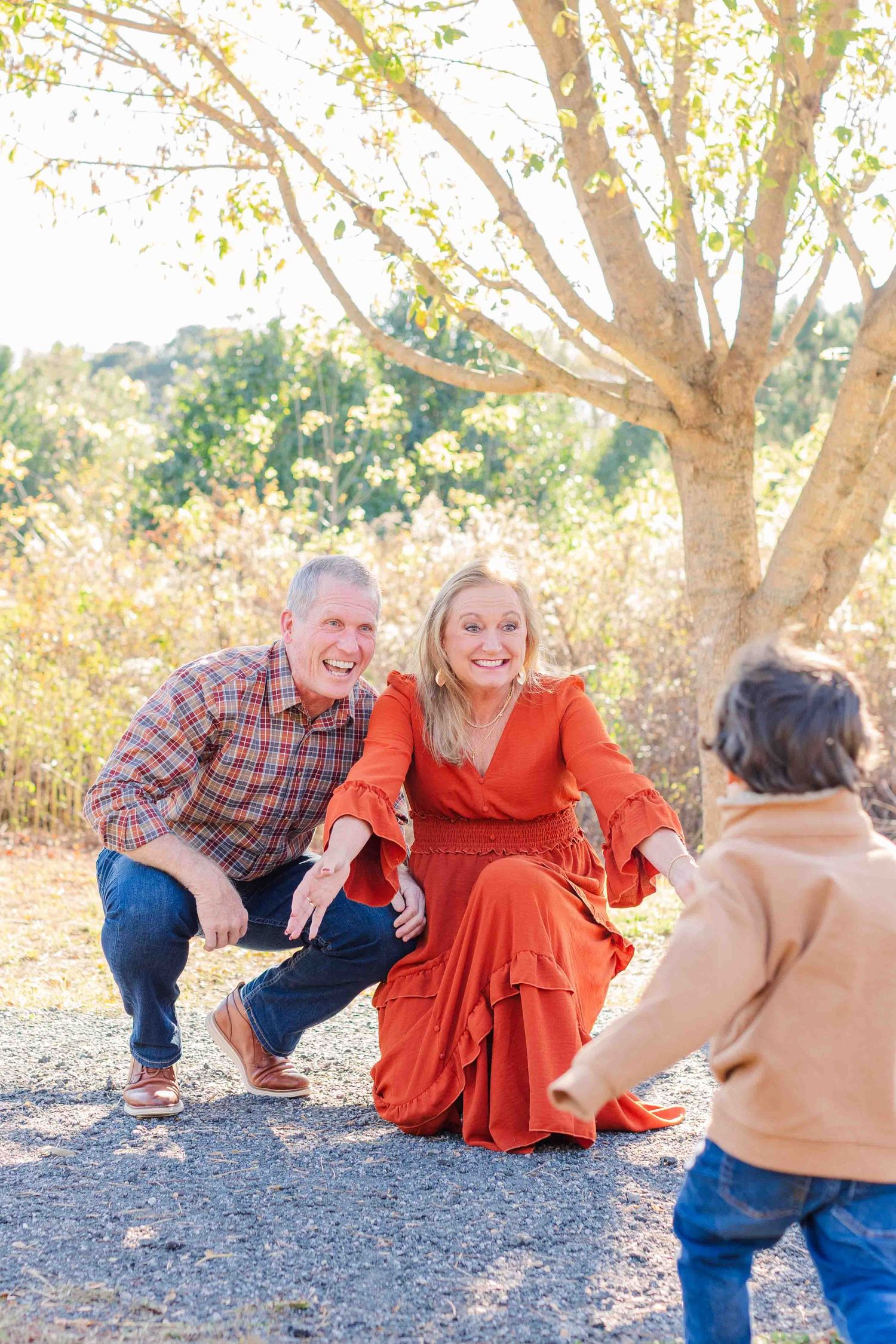 Smiling grandparents crouching down to welcome a young child running toward them outdoors.