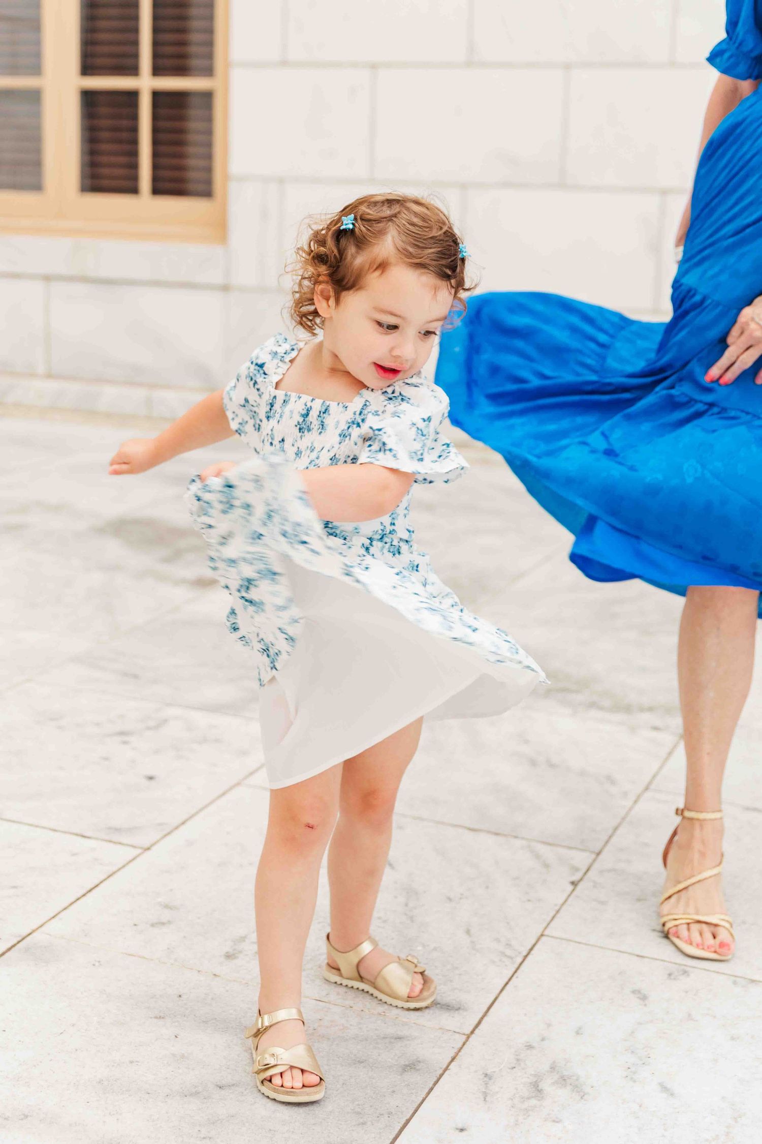 Adorable toddler girl in white floral dress twirling outdoors near marble building.