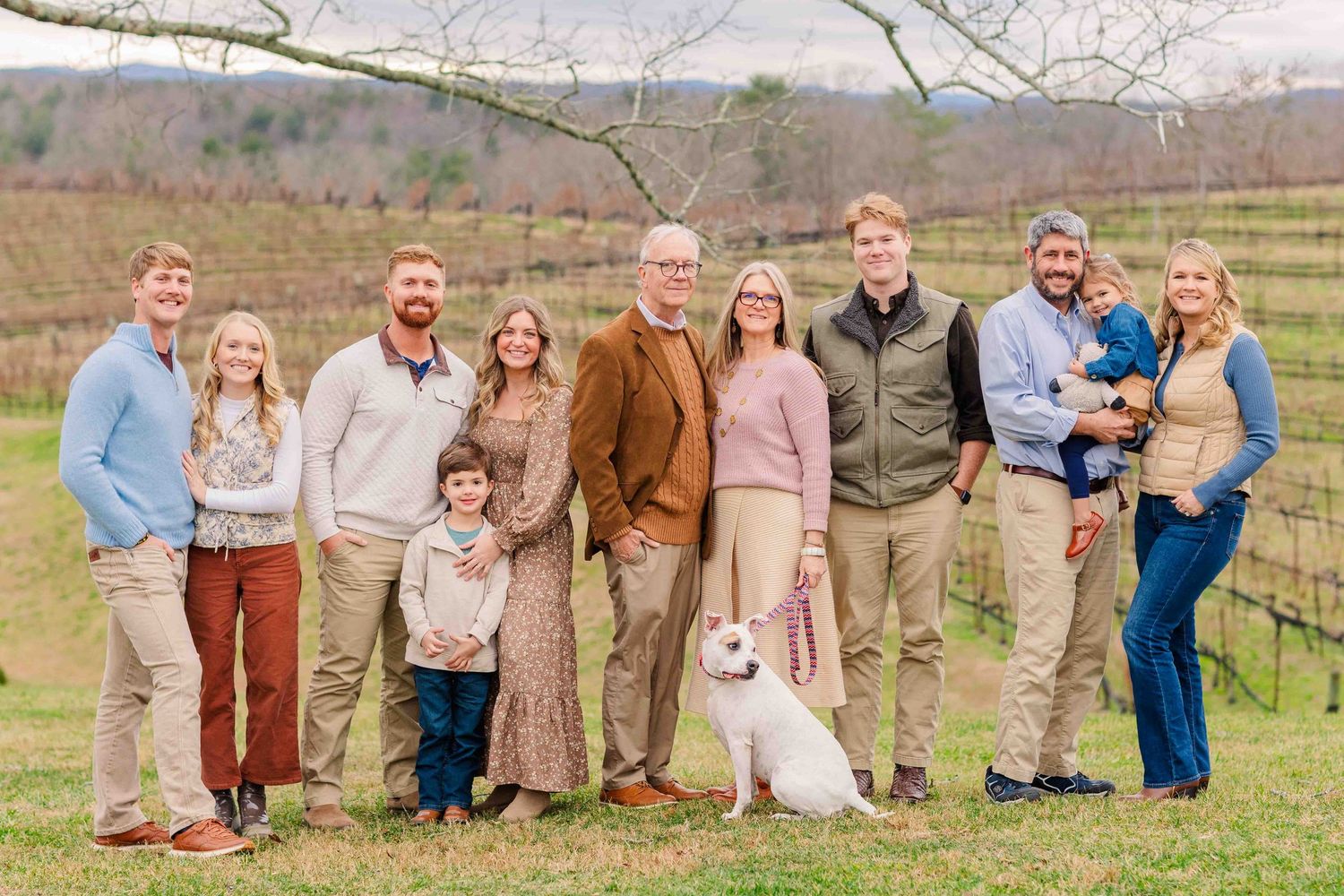 Large extended family group portrait with a white dog posing outdoors in a winter vineyard.