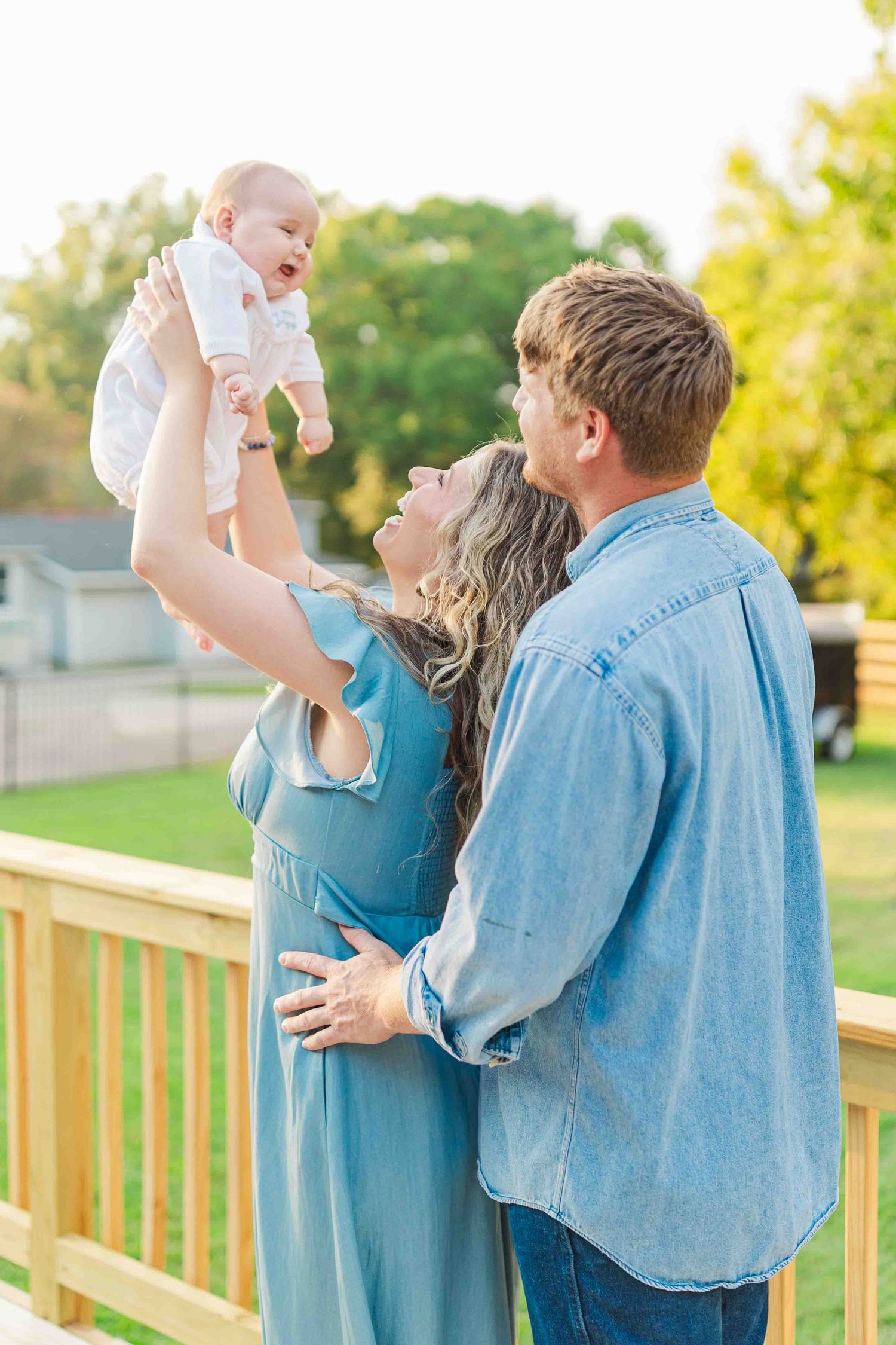 Joyful parents lifting laughing baby in the air on a sunny wooden deck in denim outfits.