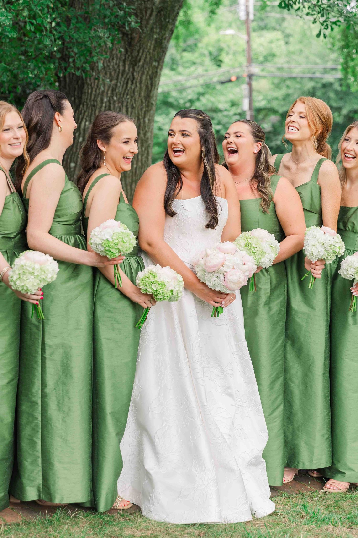 Bride in white gown laughing with bridesmaids in green dresses holding white floral bouquets outdoors.