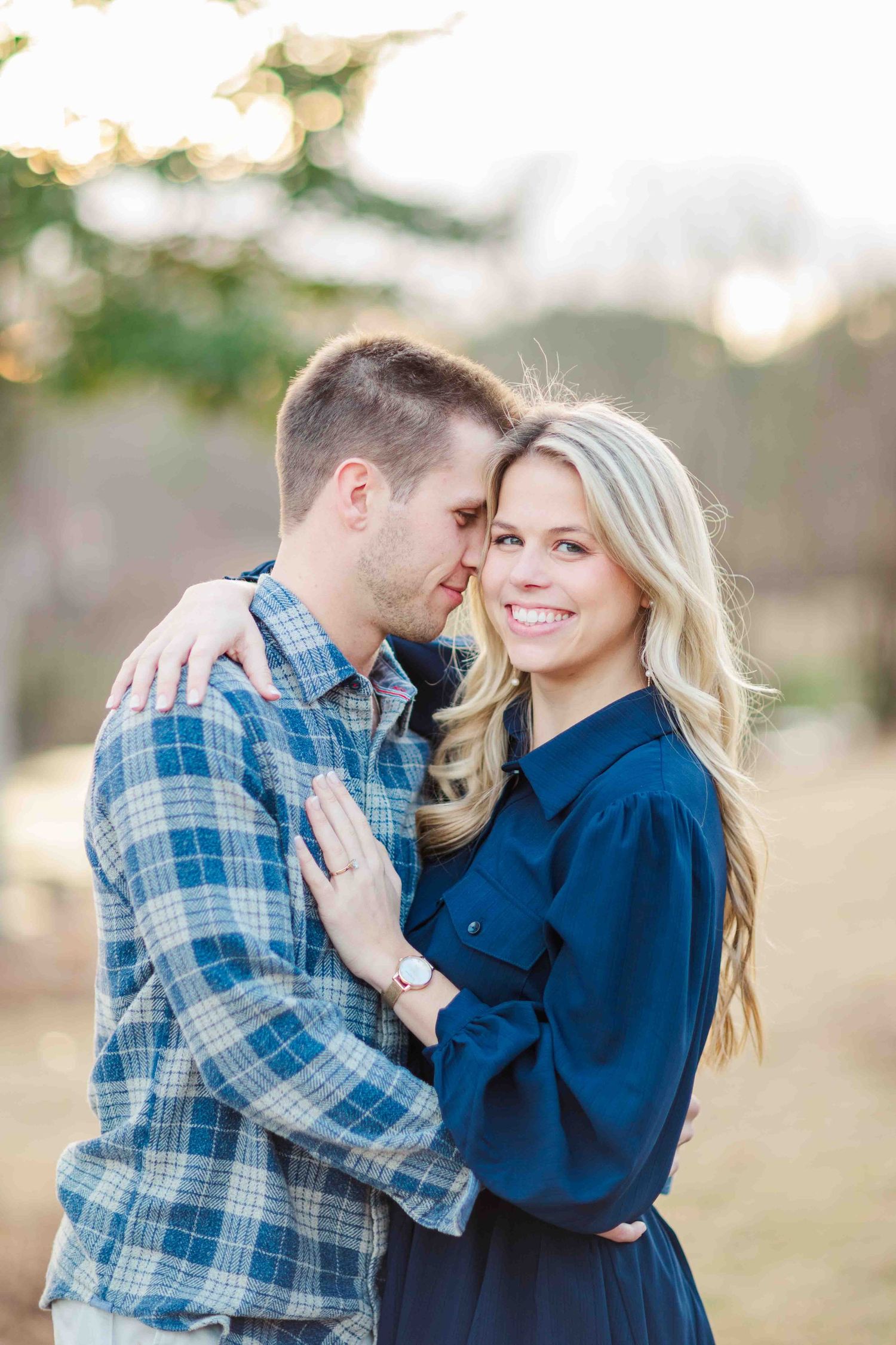 Smiling blonde woman in navy dress embracing man in plaid shirt in park.