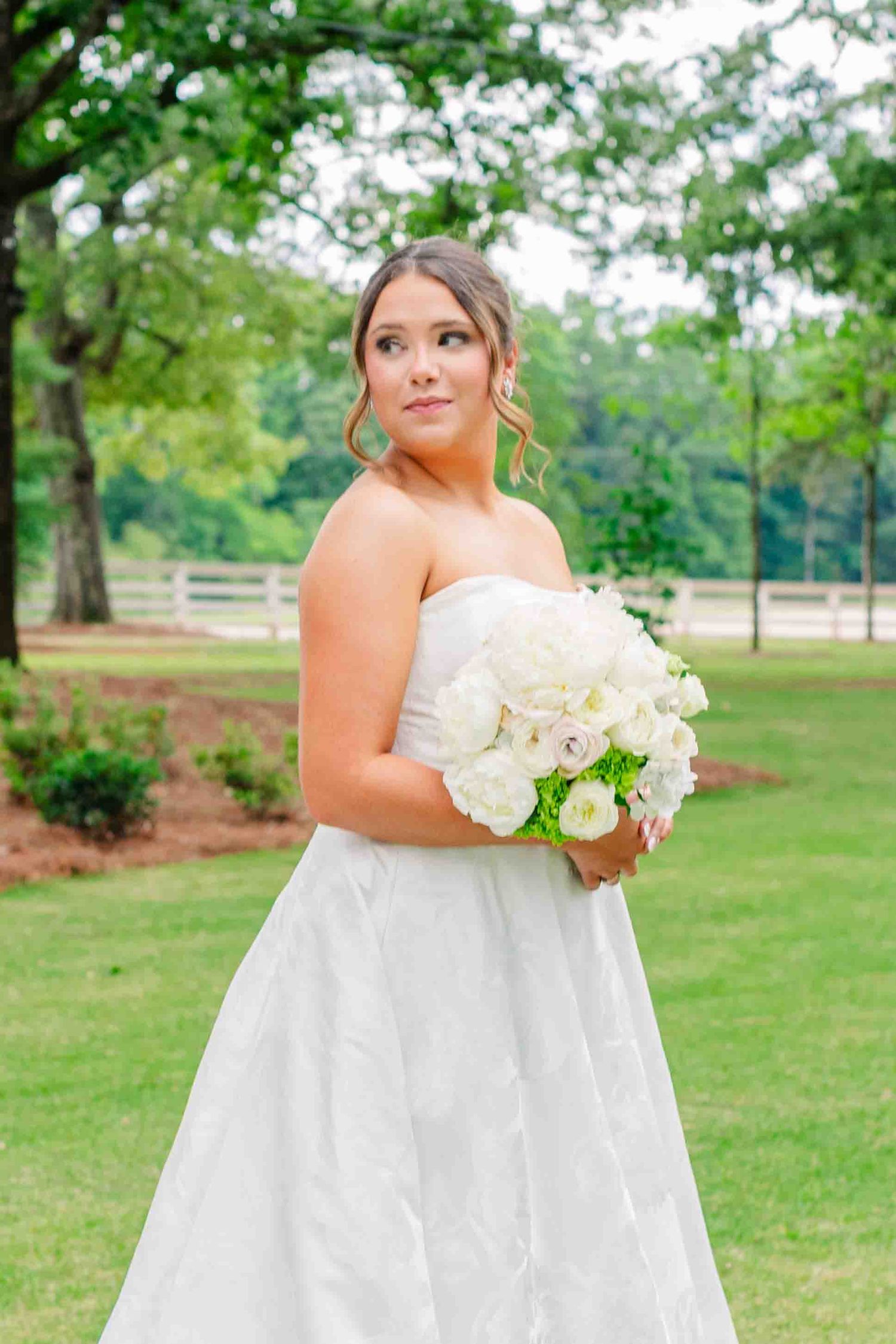 Bride in strapless white gown holding white bouquet posing outdoors in green garden.
