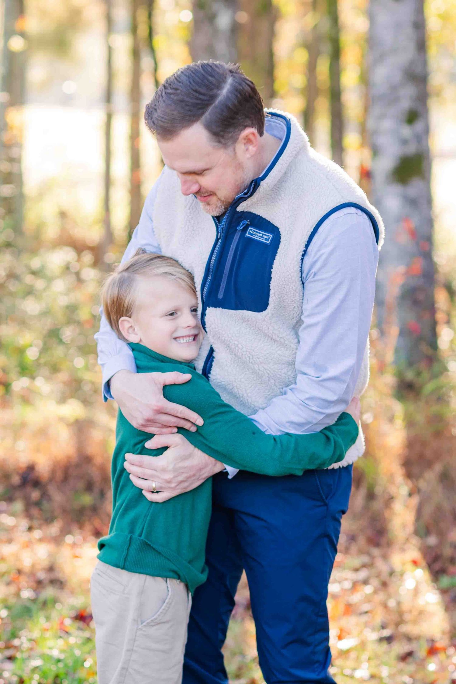 Father hugging his young smiling son in a sunlit autumn forest setting.
