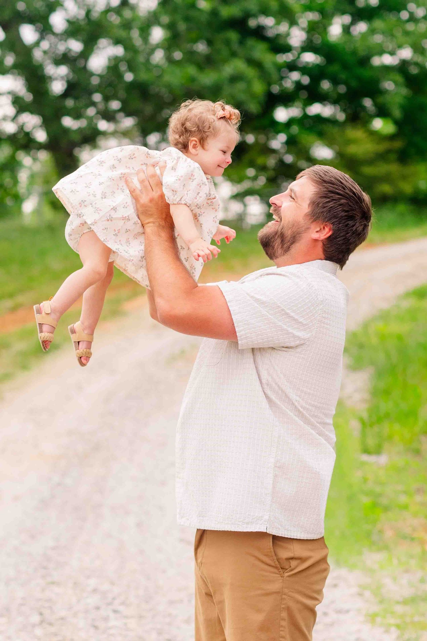Joyful father lifting smiling toddler daughter into the air on a sunny outdoor gravel path.