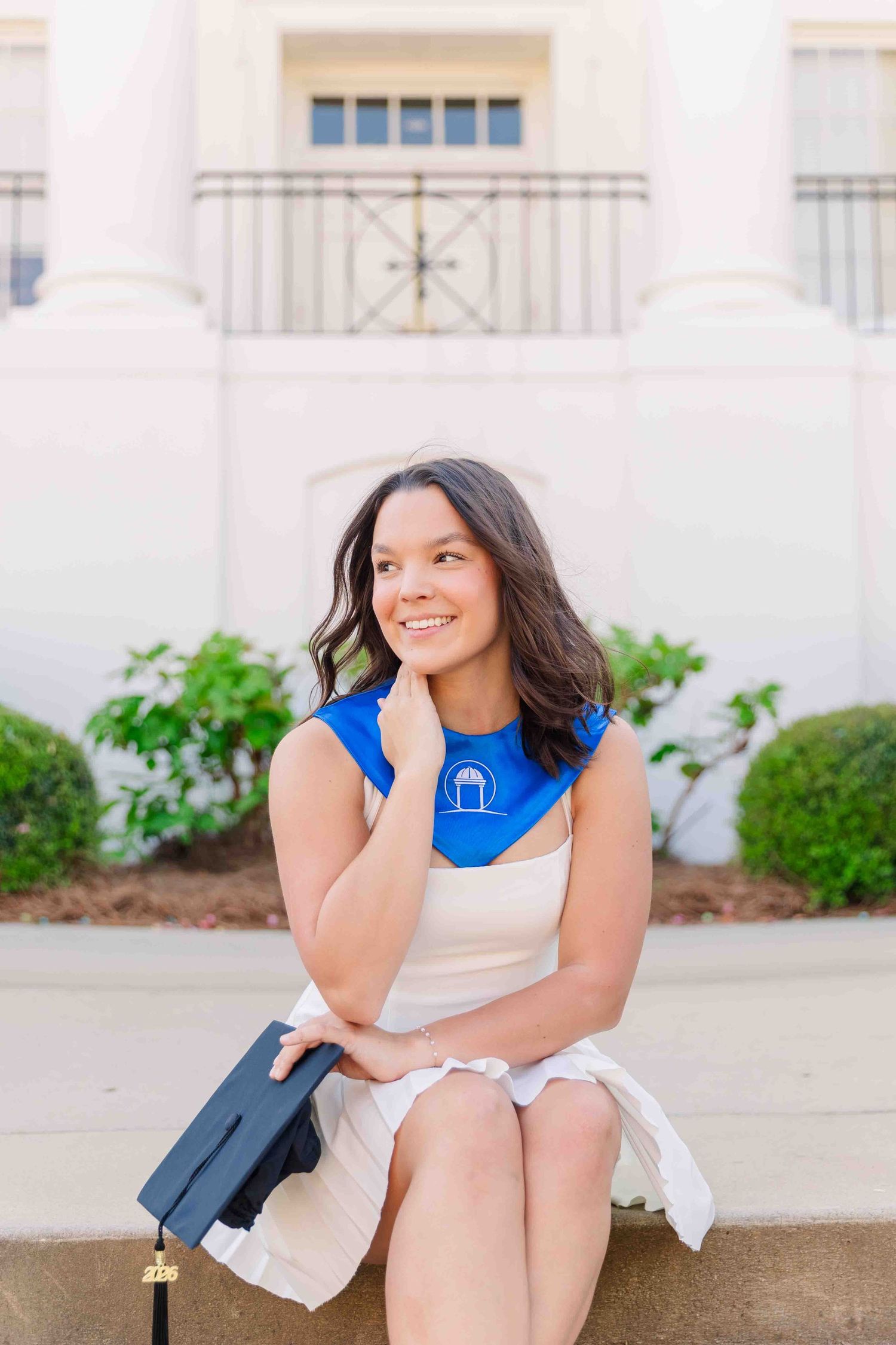 Brunette woman in white dress with blue graduation stole sits smiling holding black mortarboard cap.