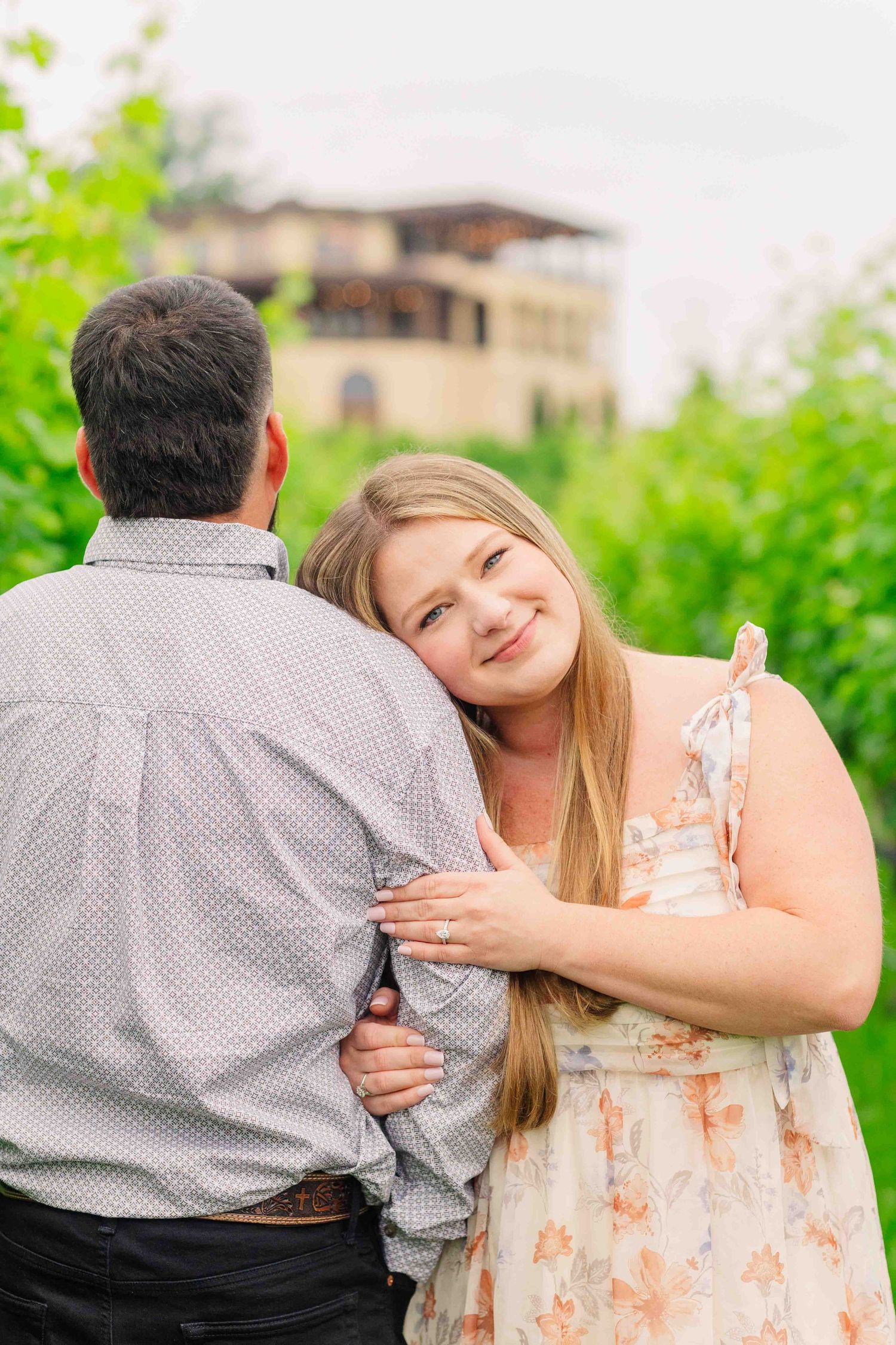 Smiling woman in floral dress hugging man from behind in lush green garden.