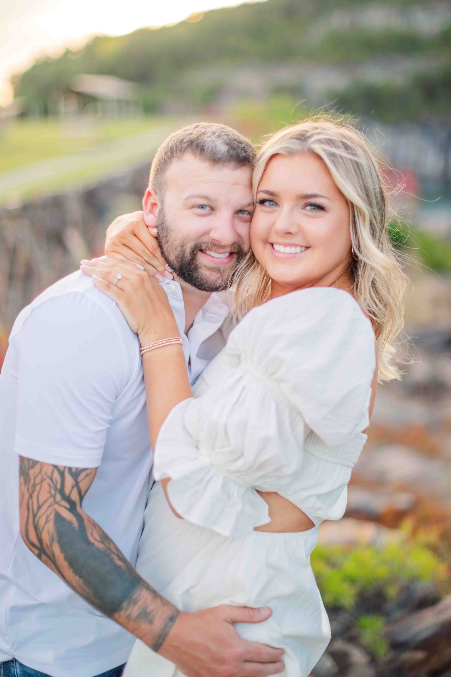 Smiling couple embracing outdoors at sunset, woman in white dress with man in white shirt showing tattoos.