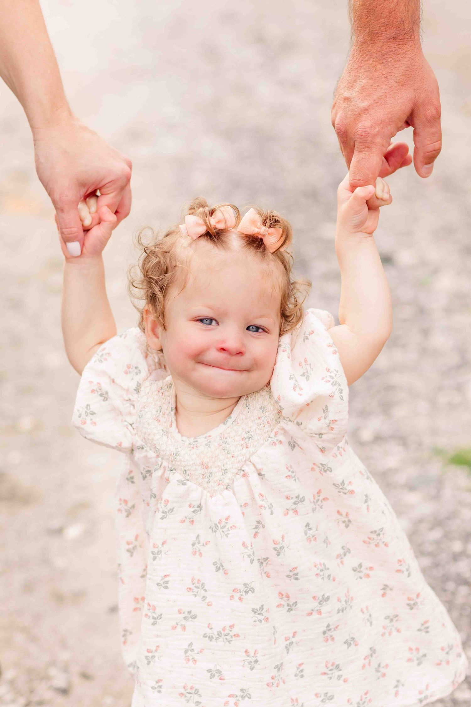 Adorable curly-haired toddler girl in floral dress holding parents hands while smiling outdoors.