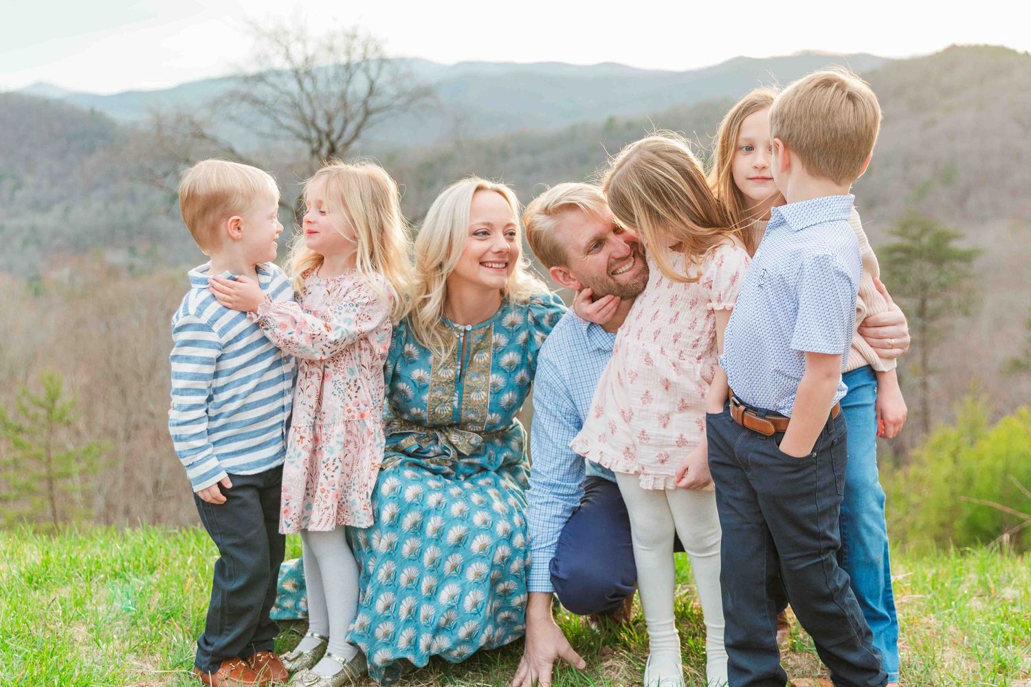 Joyful family with five young children laughing and hugging outdoors on a green hillside together.