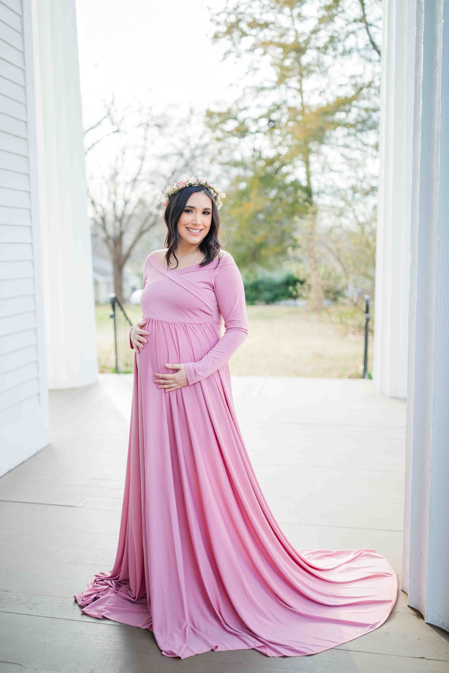 Elegant pregnant woman in flowing pink maxi dress with floral crown on porch.