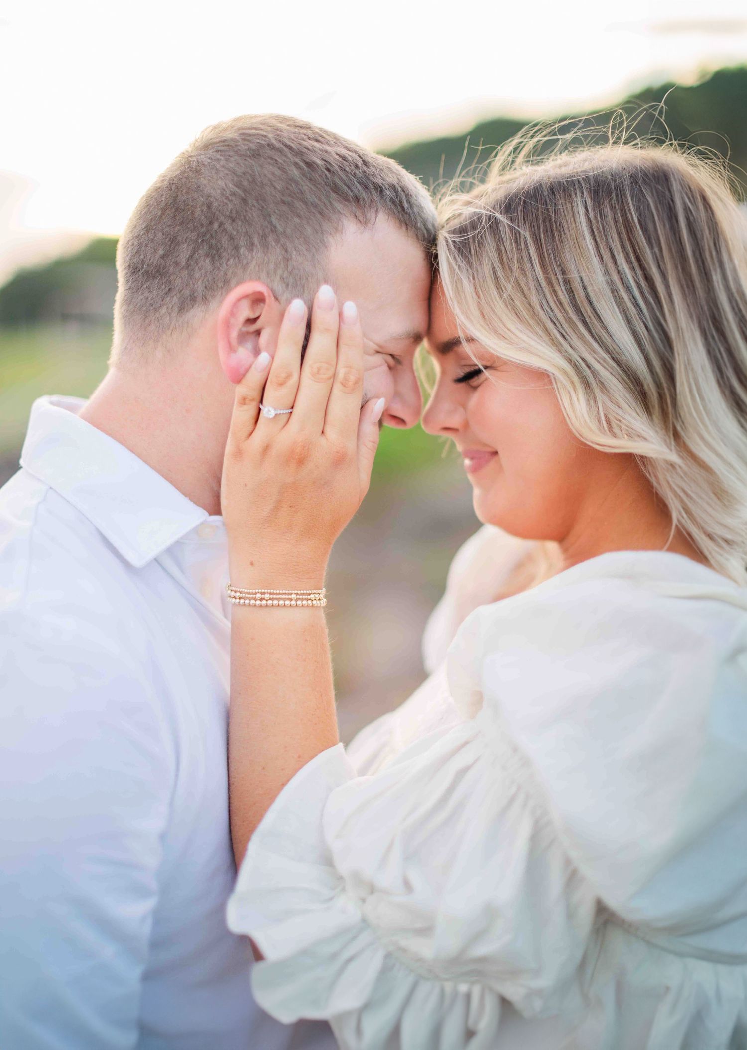 Romantic close-up of engaged couple touching foreheads tenderly, woman showing diamond engagement ring at sunset.
