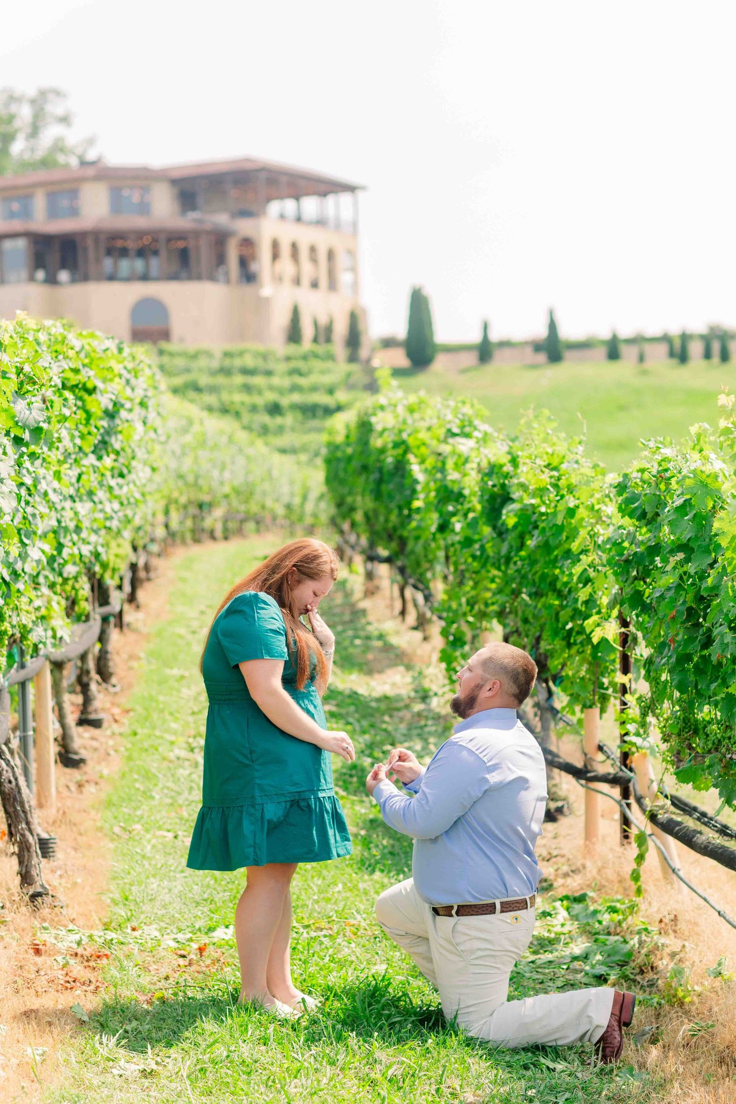Man proposing on one knee to woman in green dress among lush vineyard rows with winery building in background.