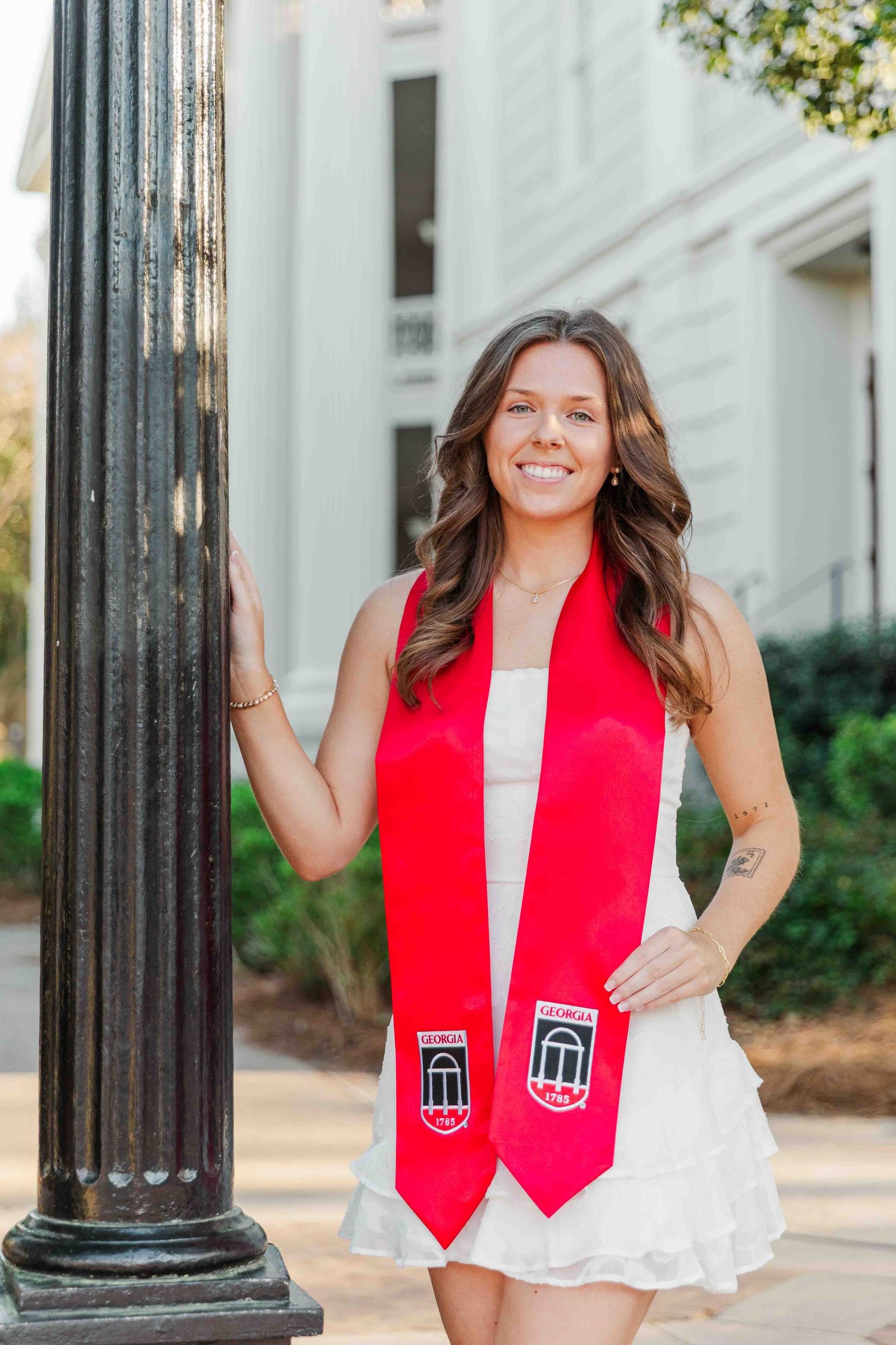 Smiling graduate wearing red graduation stole over white dress, posing by black lamppost outdoors.