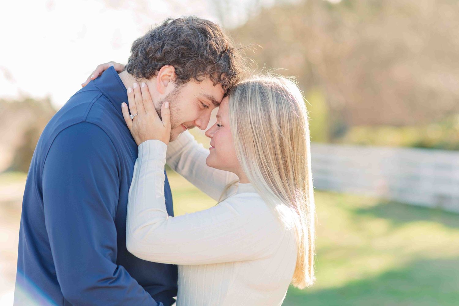 Romantic couple nearly kissing in golden sunlight outdoors, woman in cream sweater, man in navy top.