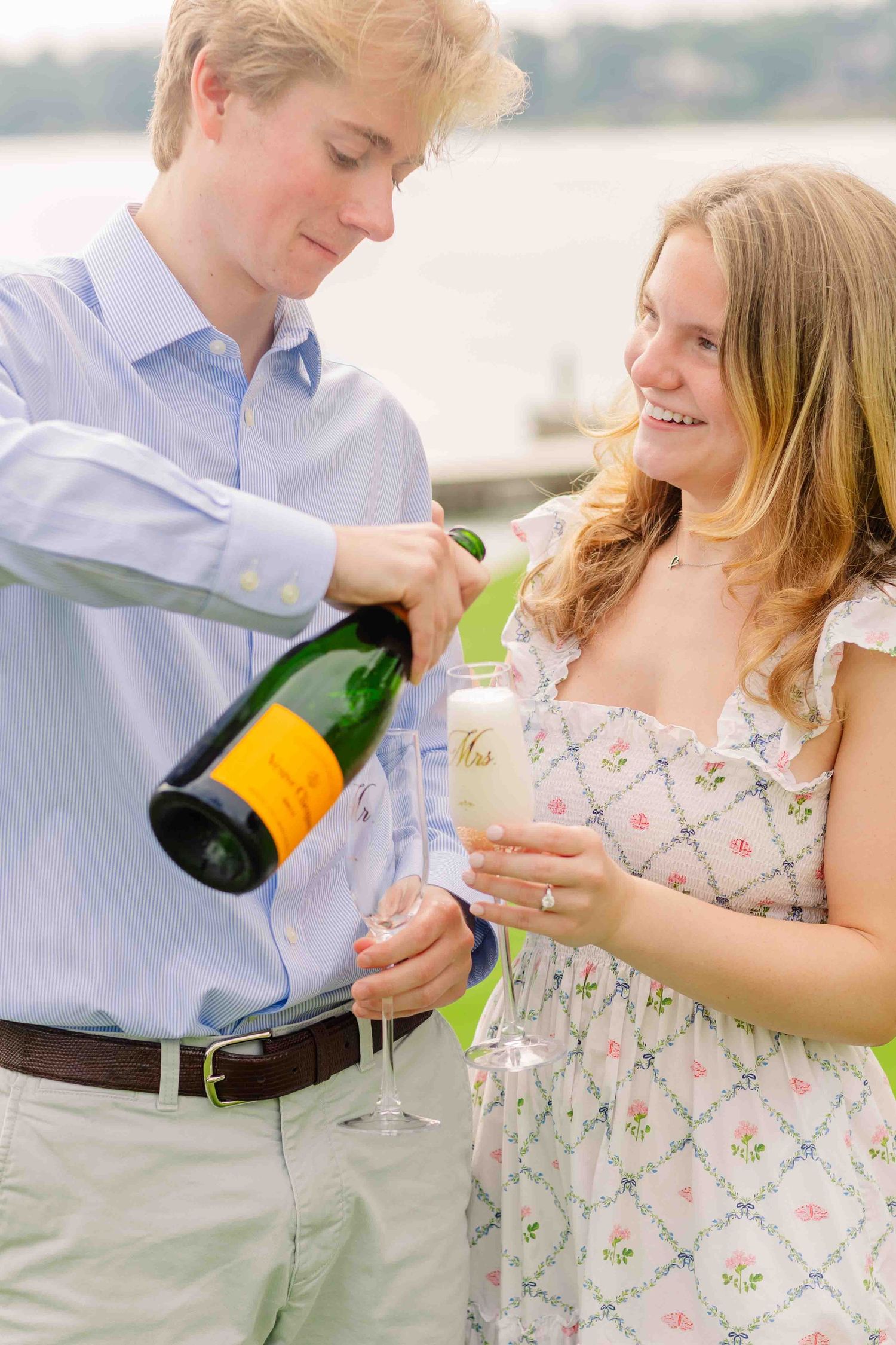 Couple celebrating engagement with champagne by the water, man pouring Veuve Clicquot into glasses.