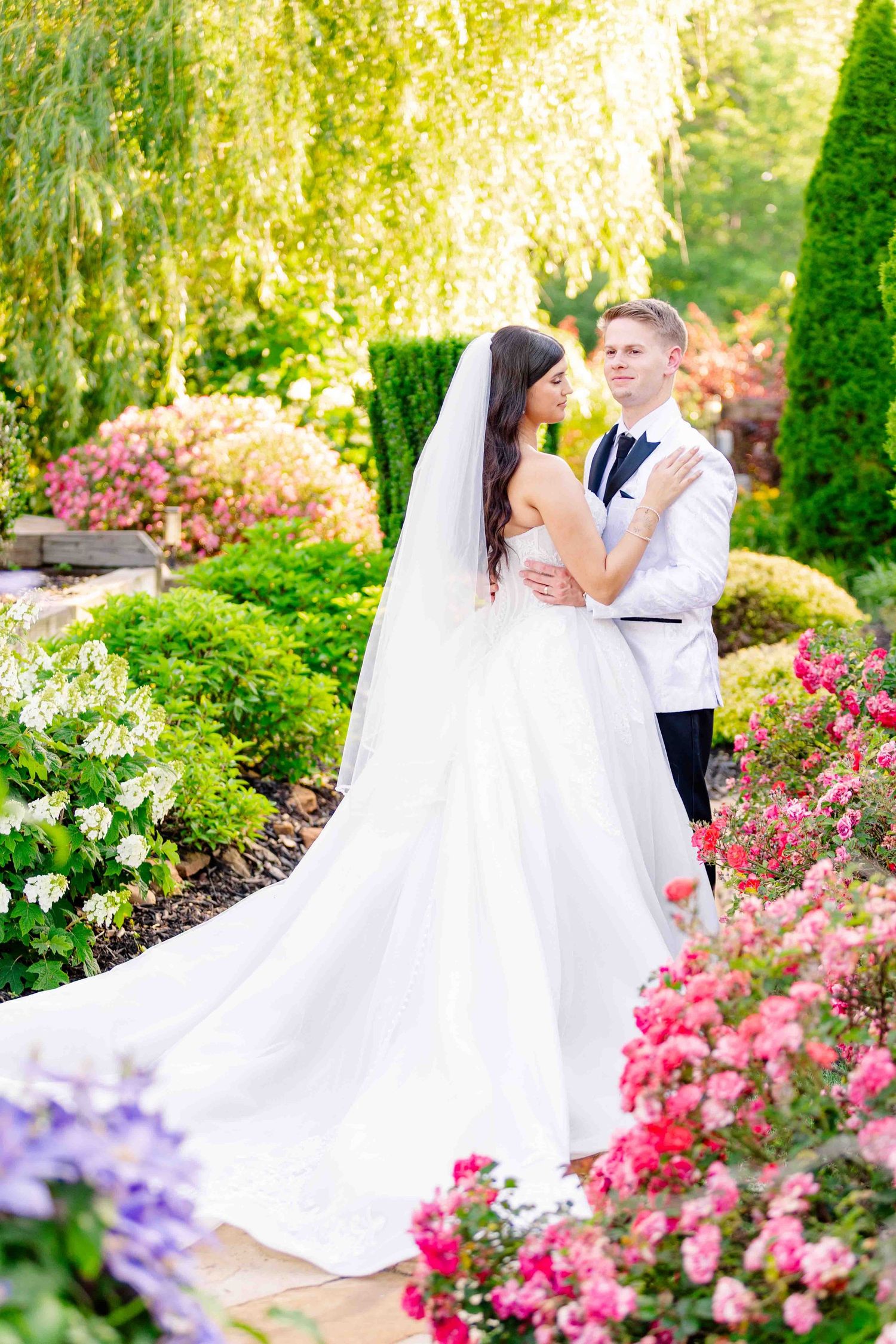 Bride and groom embrace in a stunning garden filled with colorful roses and lush greenery under golden sunlight.
