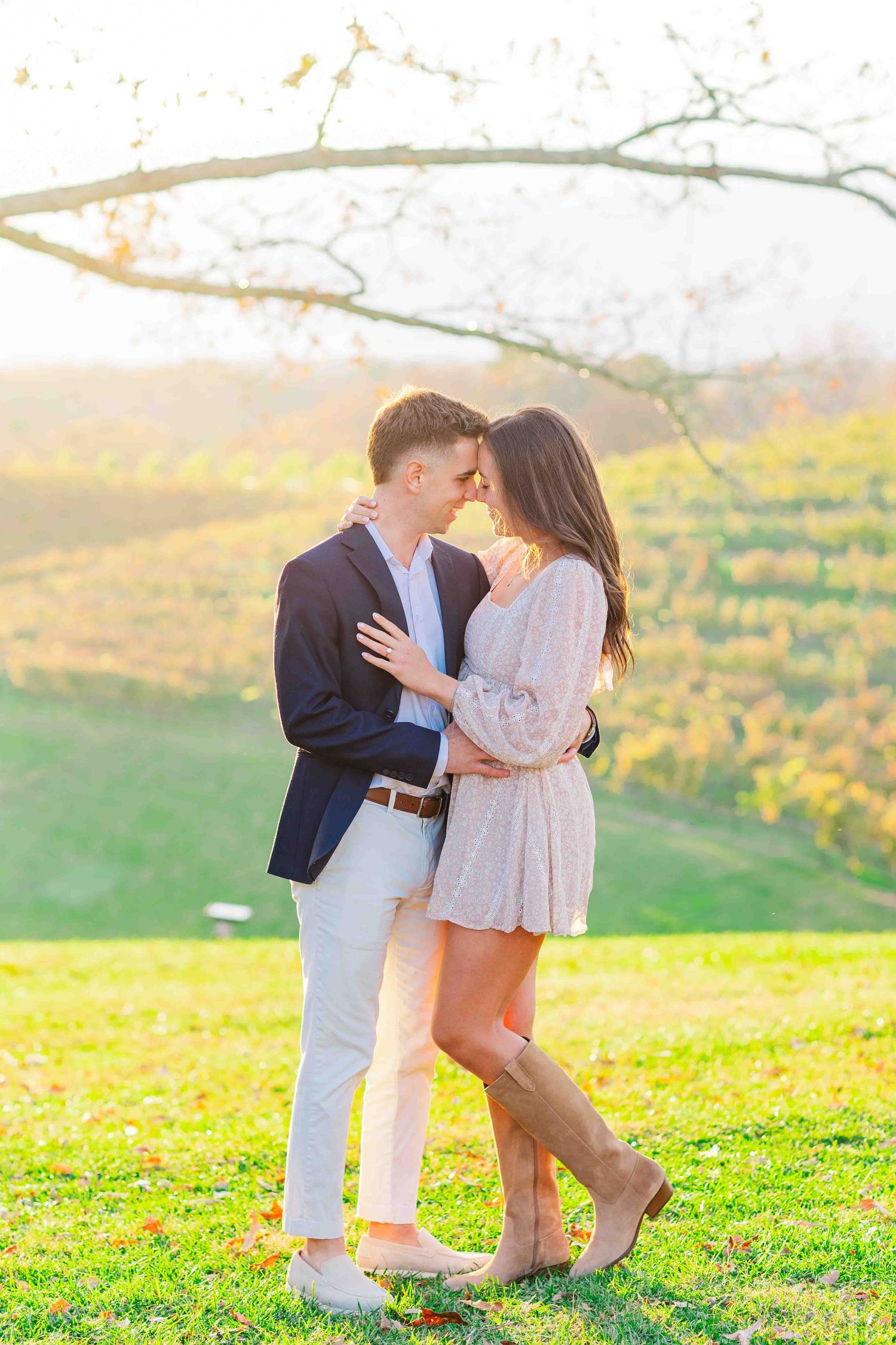 Couple embracing tenderly at golden hour in a scenic vineyard, woman in short dress wearing cowboy boots.