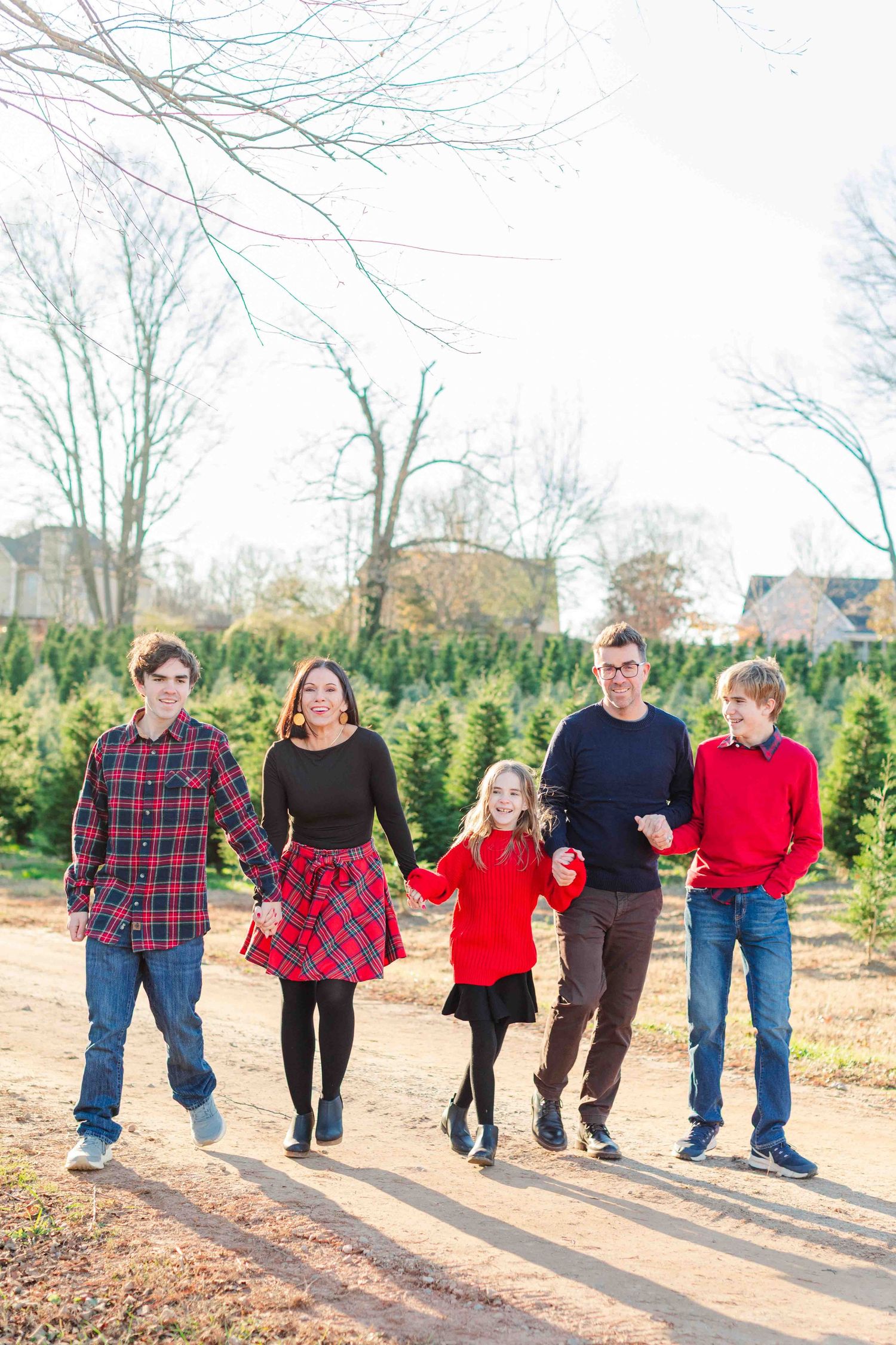 Family of five walking together at a Christmas tree farm, wearing red and plaid holiday outfits.