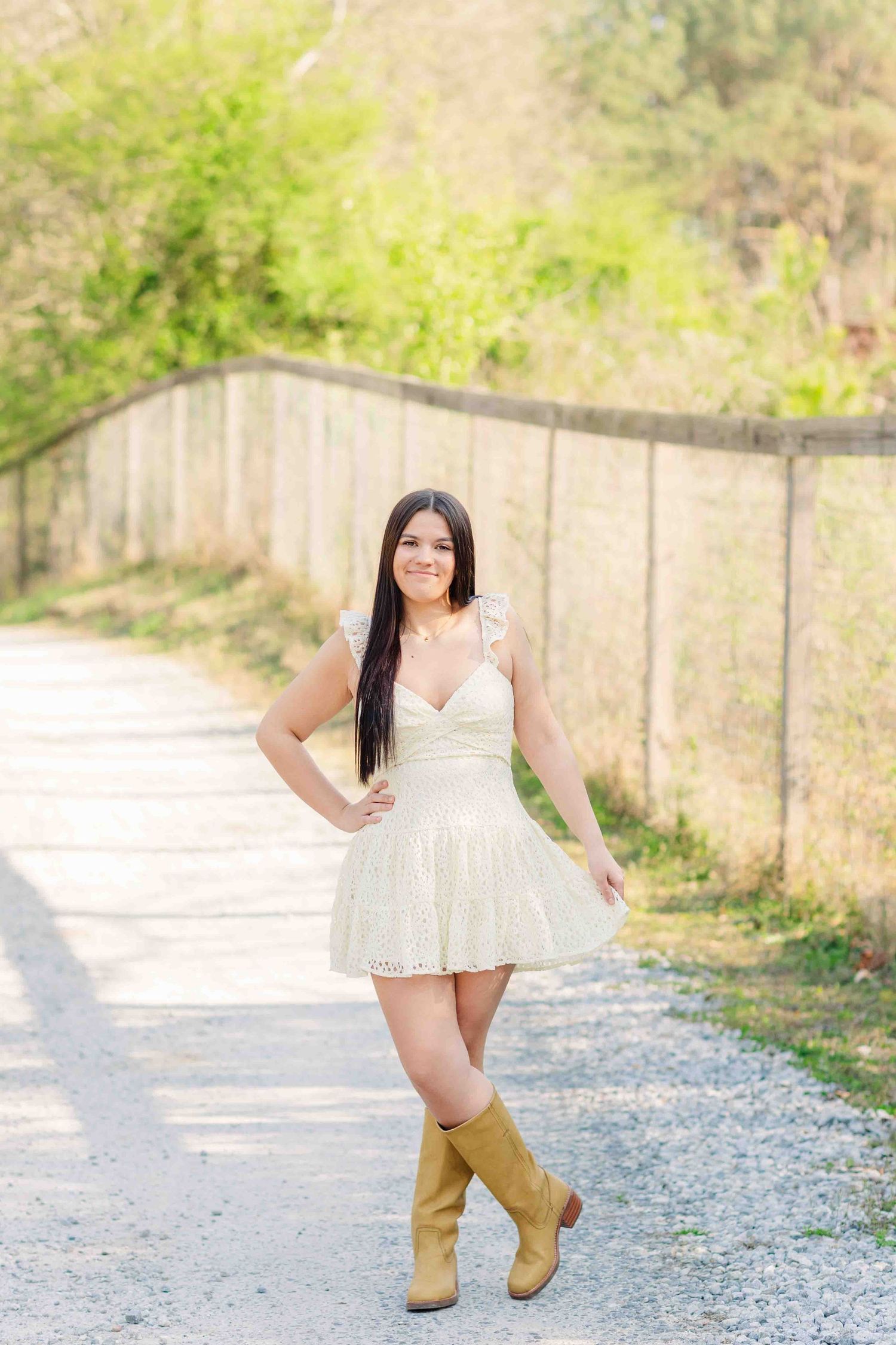Smiling brunette woman in cream dress and tall boots posing on sunny gravel path outdoors.