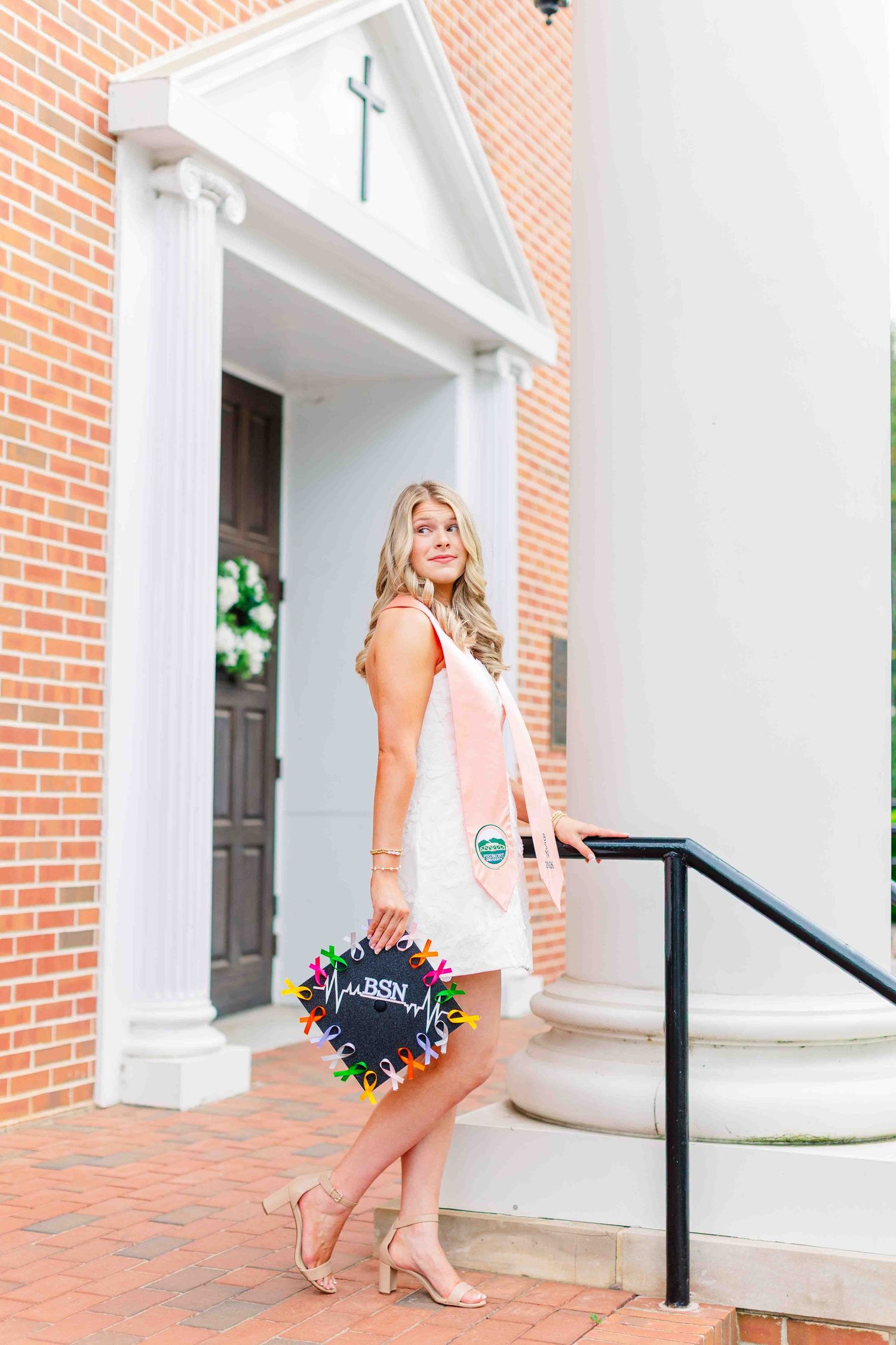 Blonde nursing graduate in white dress with pink stole holding decorated BSN cap on brick steps.