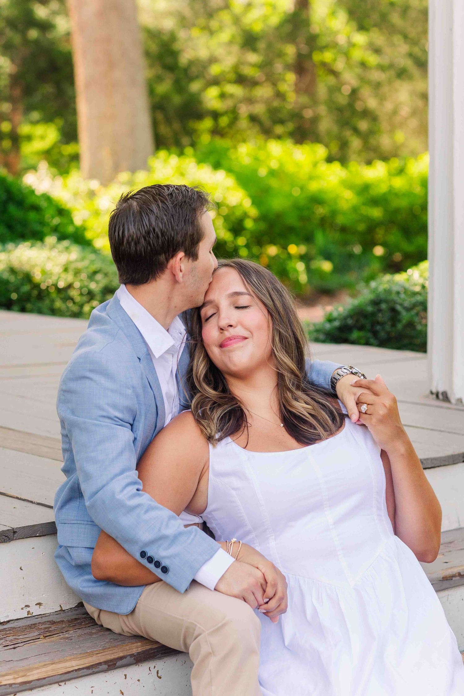 Couple sitting on steps, man kissing womans temple as she smiles with eyes closed in lush garden setting.