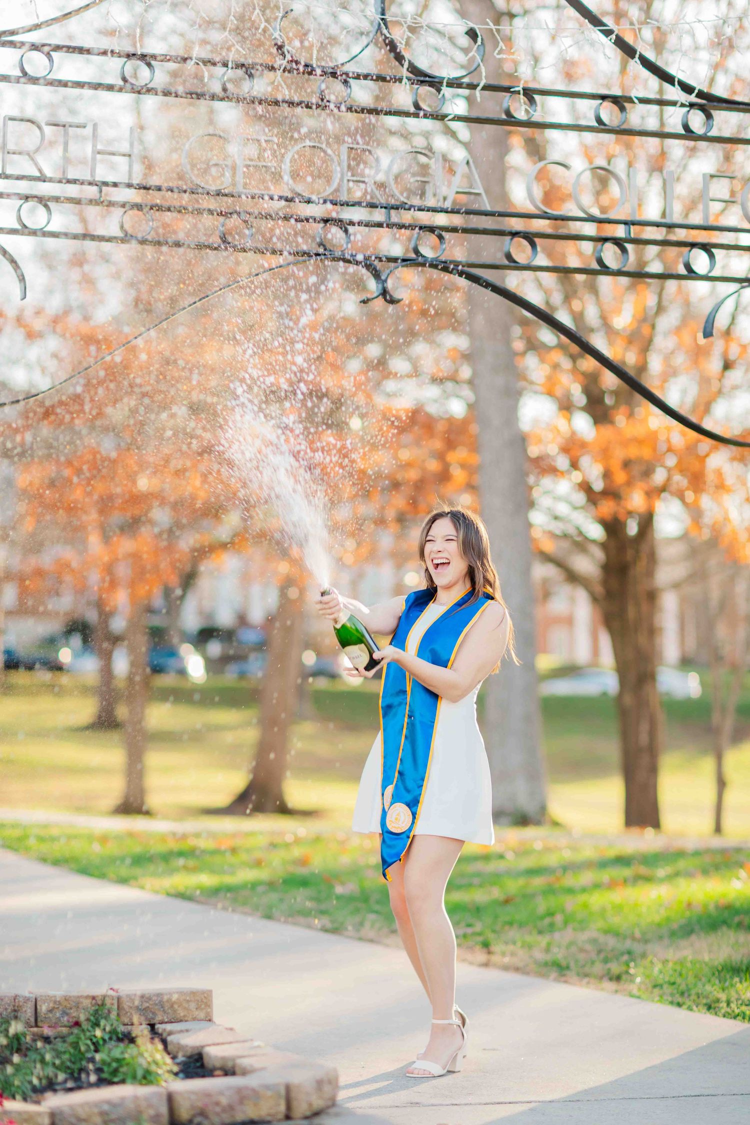 A joyful graduate in a white dress and blue stole pops champagne outside North Georgia College.