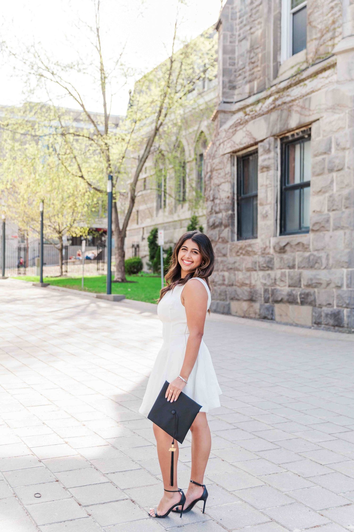 Woman in white dress holding graduation cap smiles outdoors near stone building on sunny day.