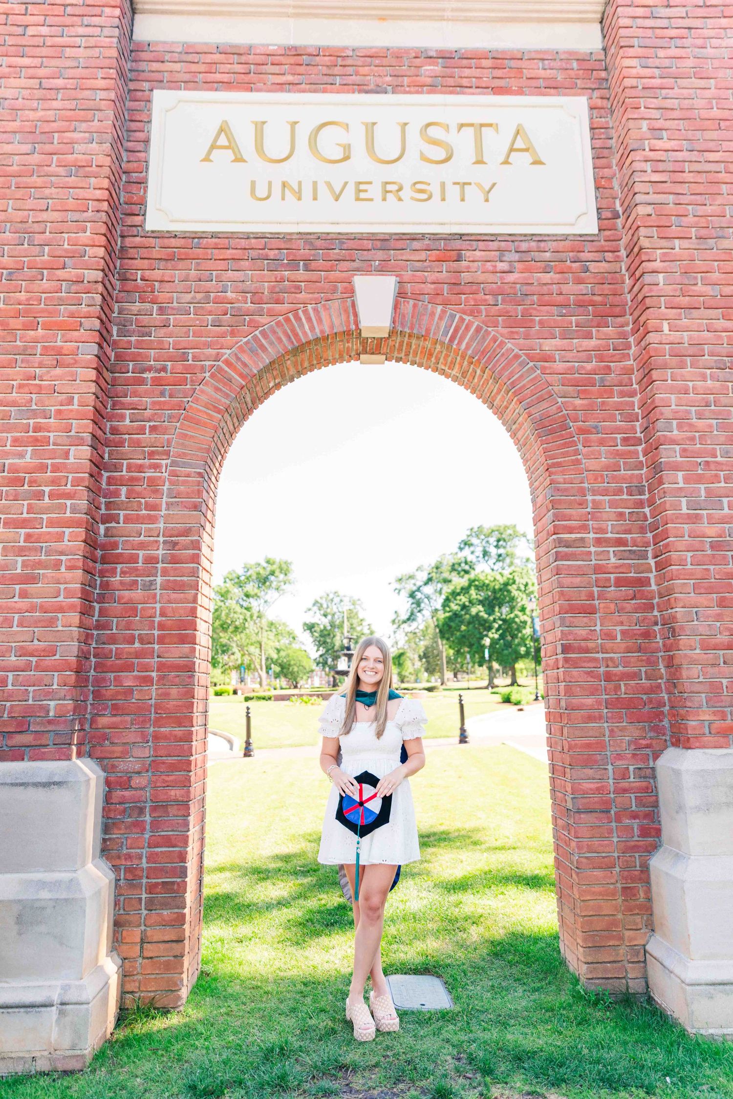 Blonde graduate in white dress holds cap under Augusta University brick arch gateway.
