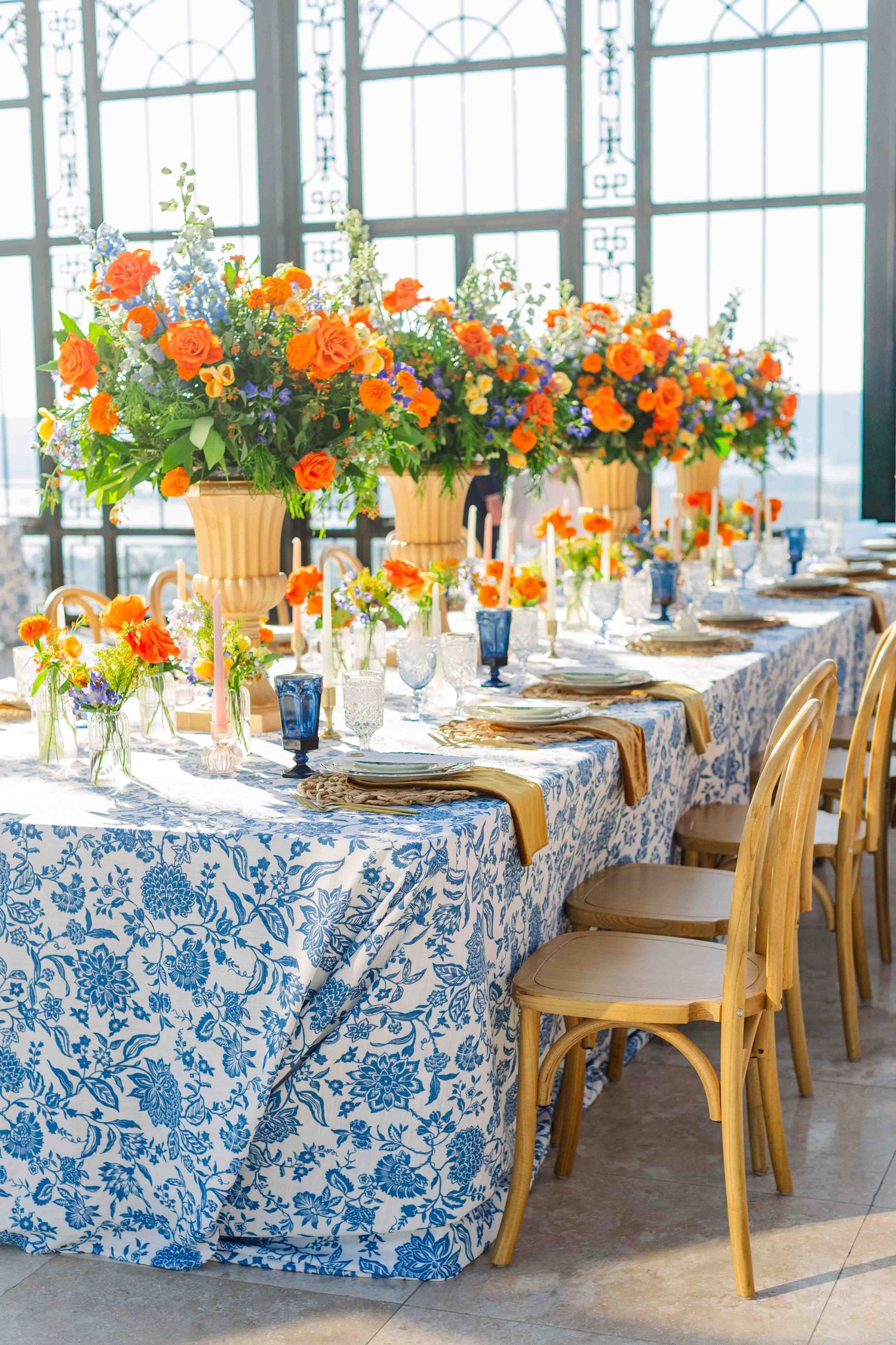 Elegant wedding reception table with blue floral linen, gold chairs, and vibrant orange flower centerpieces in a sunlit venue.