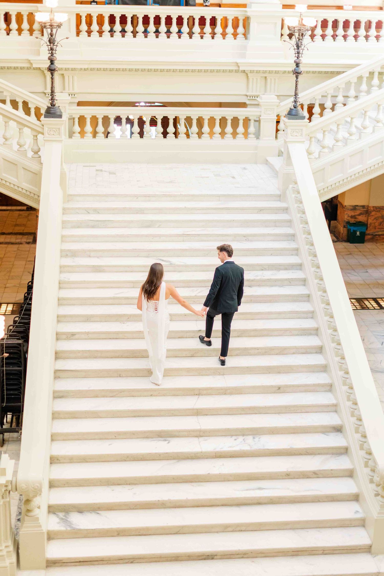 Couple holding hands walking up grand white marble staircase in elegant venue, woman in white dress, man in dark suit.
