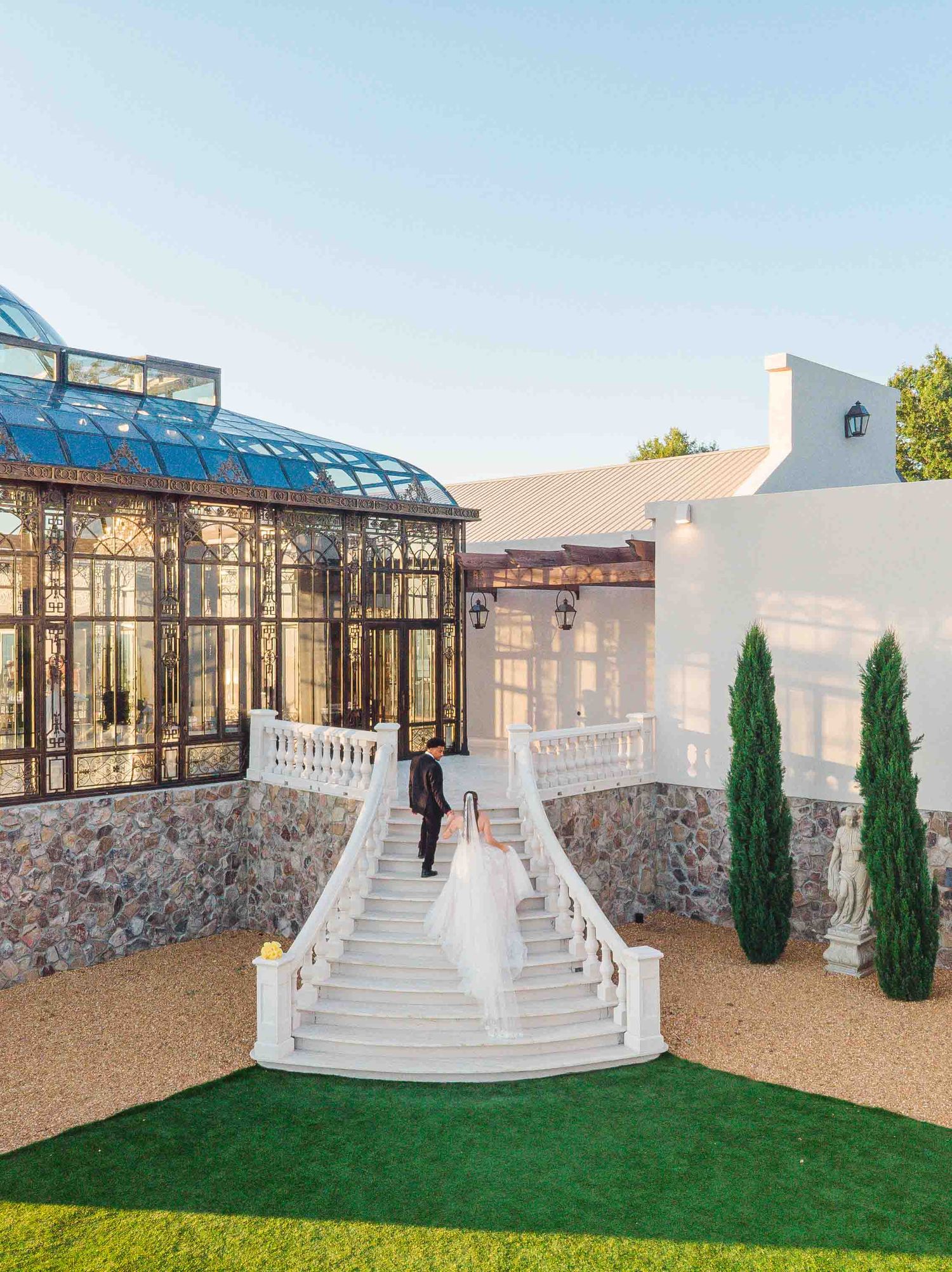 Bride and groom descend elegant white stone staircase at luxury wedding venue with glass conservatory.