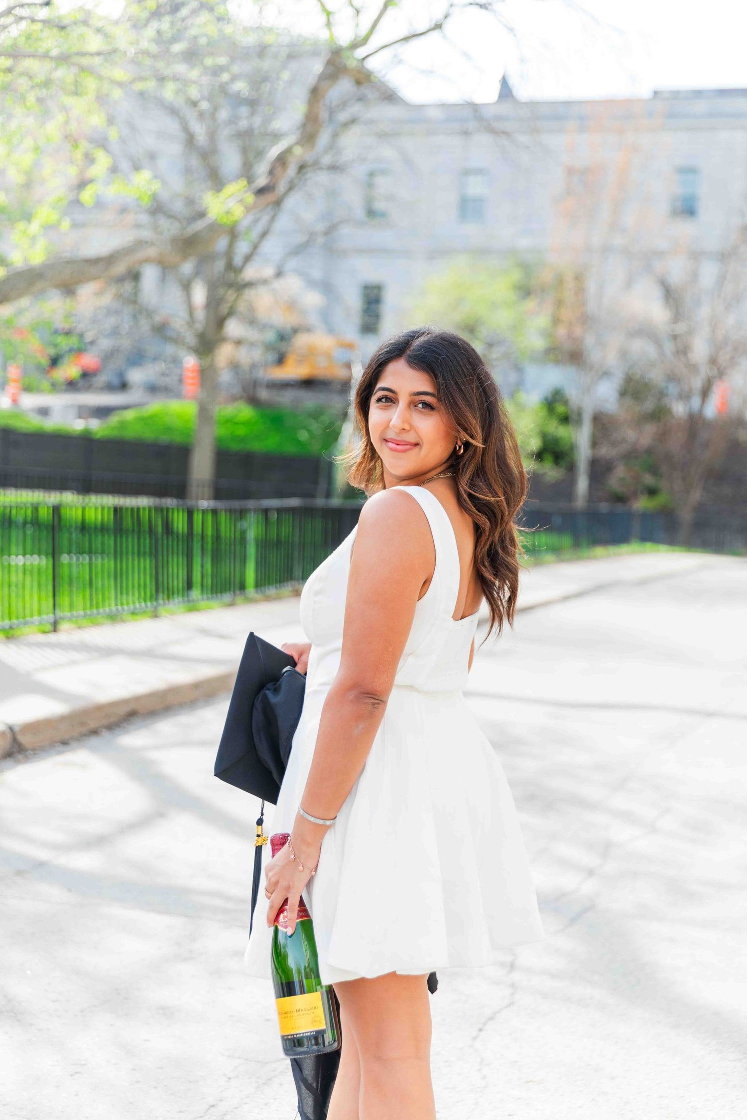 Young woman in white dress holding champagne bottle and graduation cap outdoors on a sunny day.