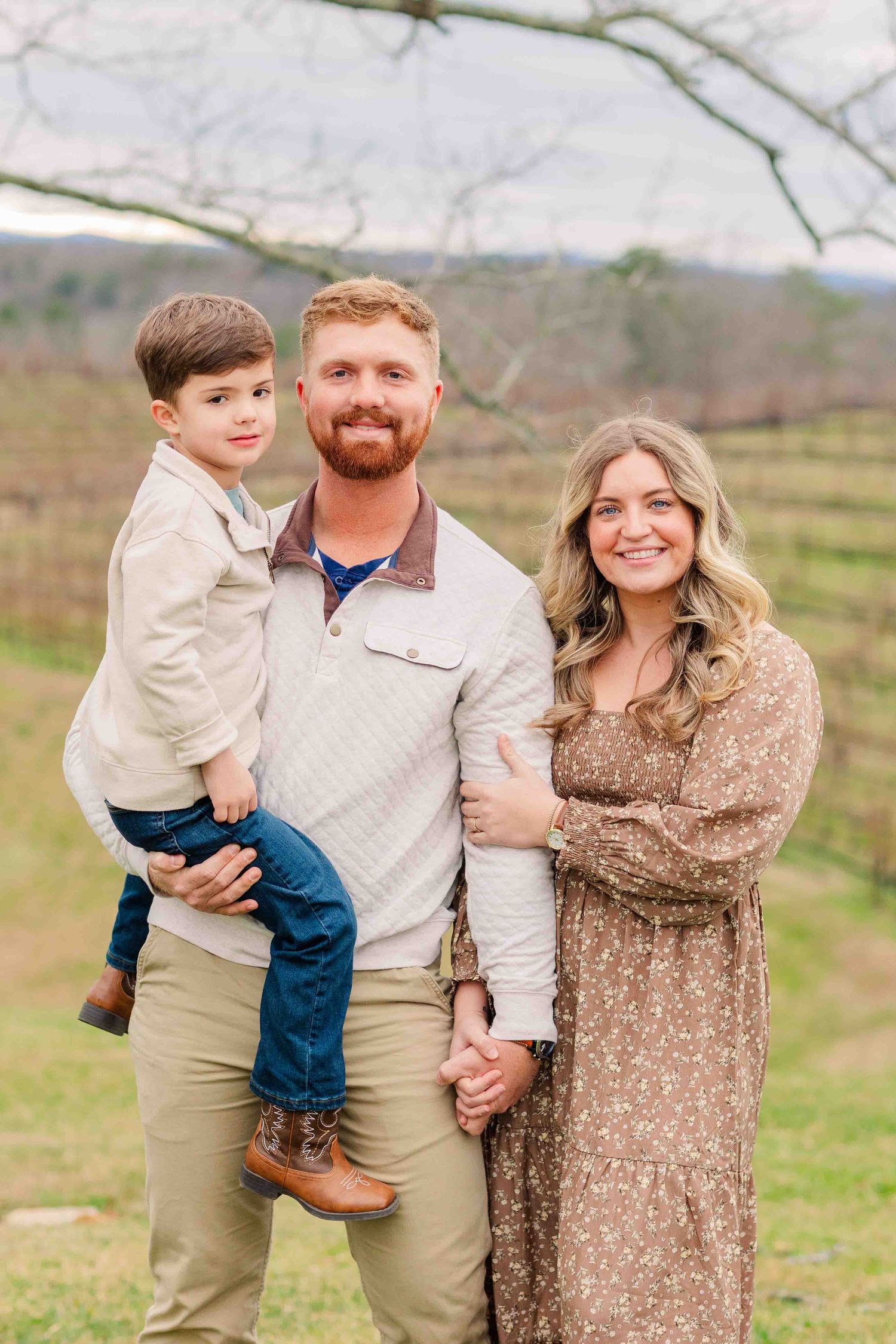 Smiling family of three posing outdoors in a vineyard, father holding young son in his arms.