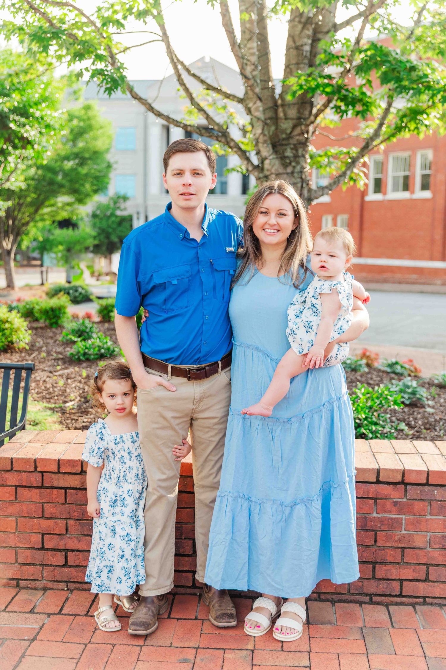 Family of four posing outdoors with two young daughters in matching blue floral dresses.