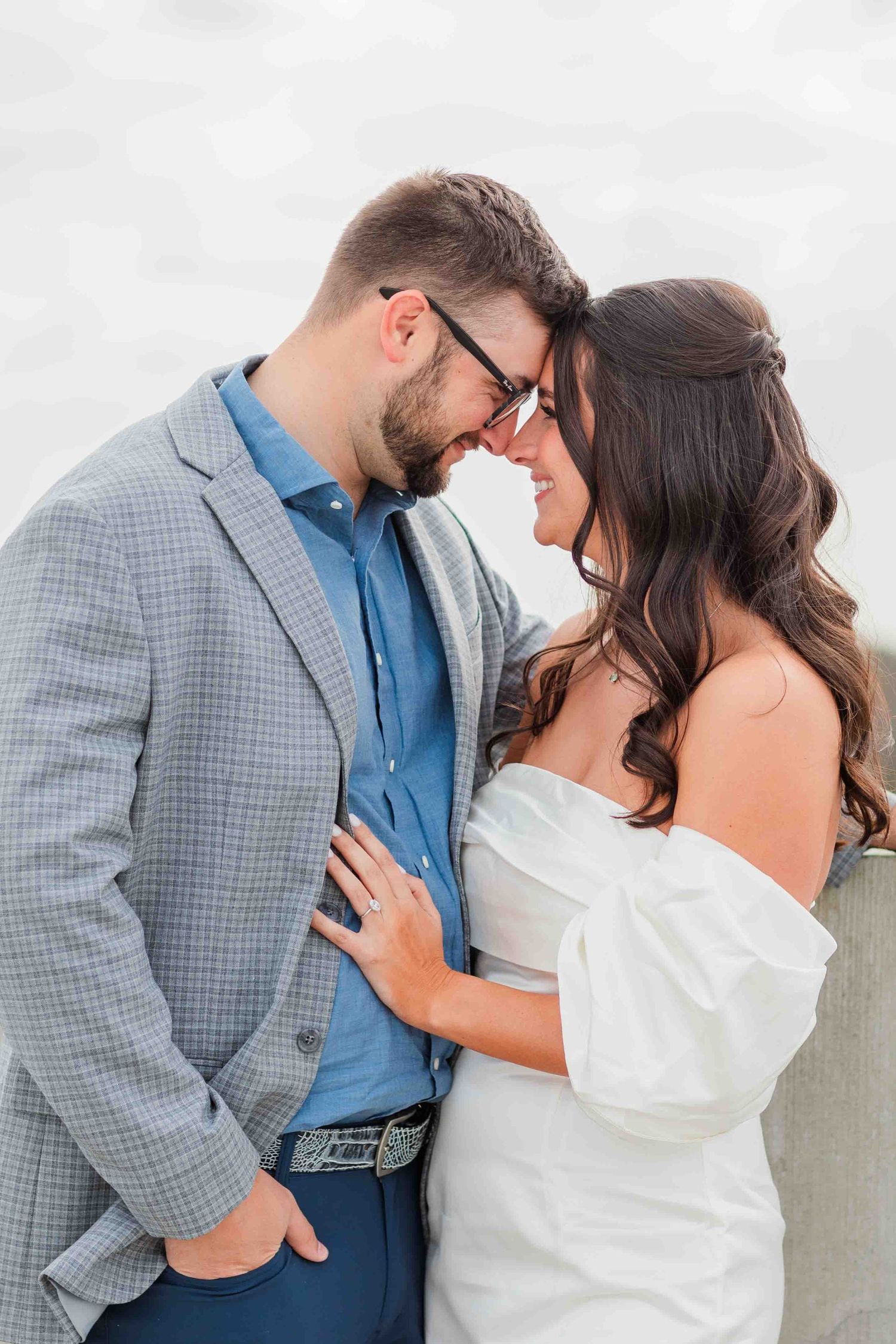 Bearded man in gray blazer and brunette woman in off-shoulder dress touching foreheads romantically.